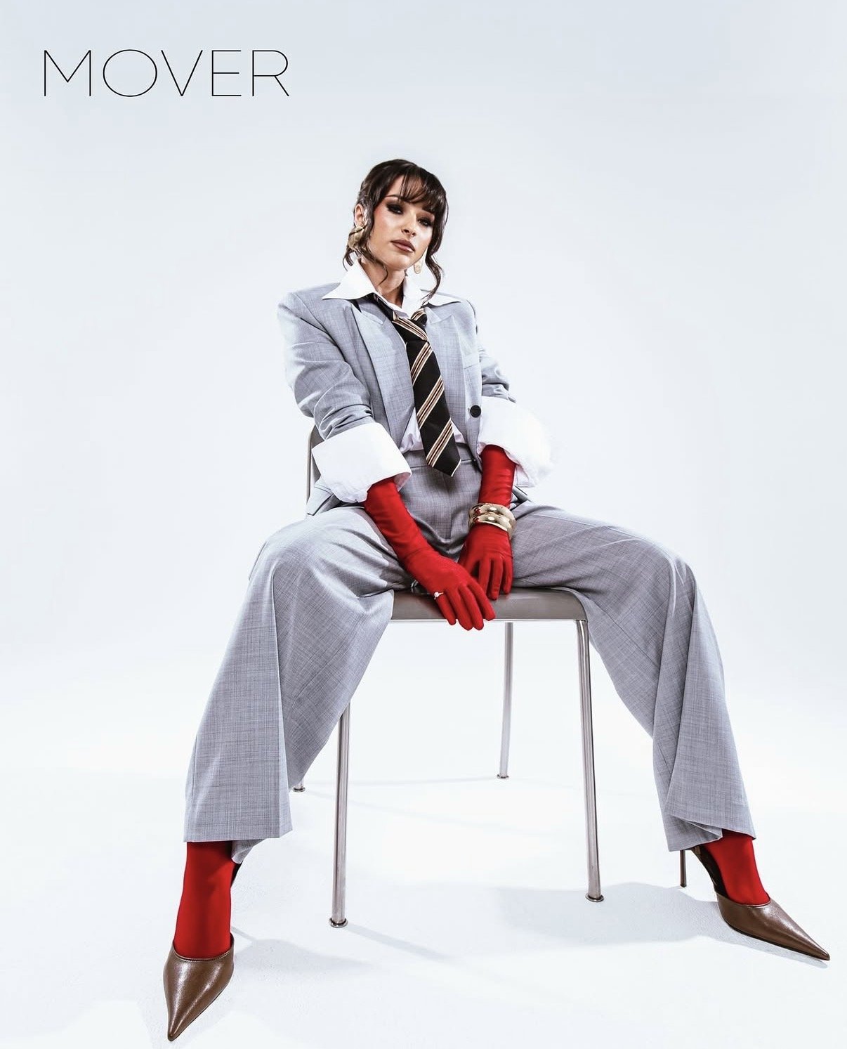 A woman dressed in a gray business suit with red gloves, red high-heeled boots, and a striped tie, sitting on a chair against a plain white background. The word "MOVER" is written in the top left corner.