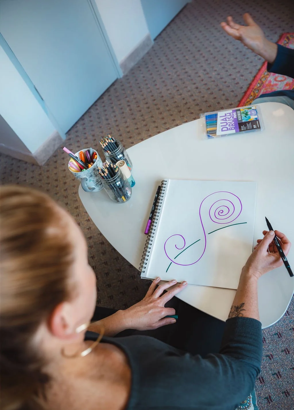 Person drawing spirals and waves on a white sketchbook with a black marker, surrounded by pens and markers on a white table.