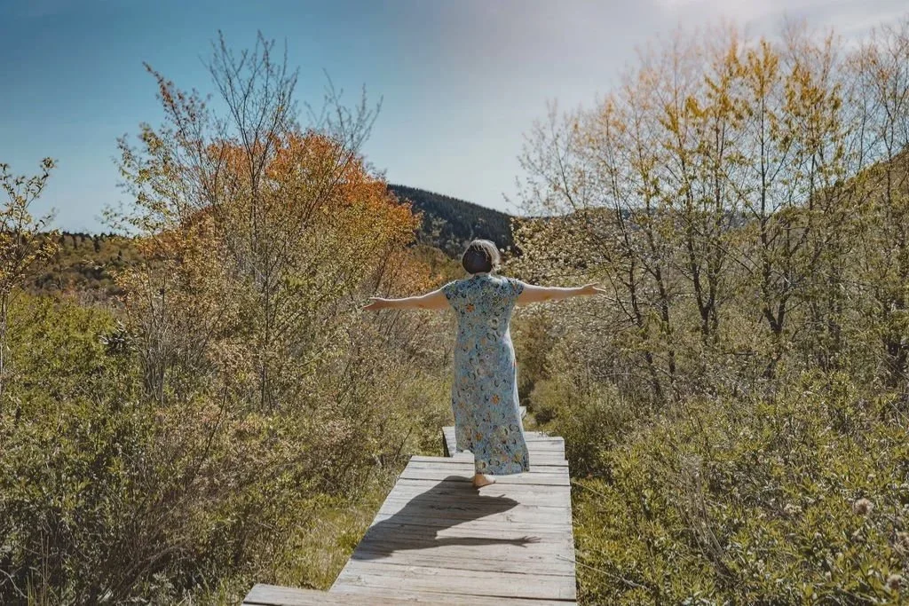 Coach and therapist Heidi Andersen in a floral dress walking through a scenic nature trail near Asheville, NC with trees and mountains in the background.