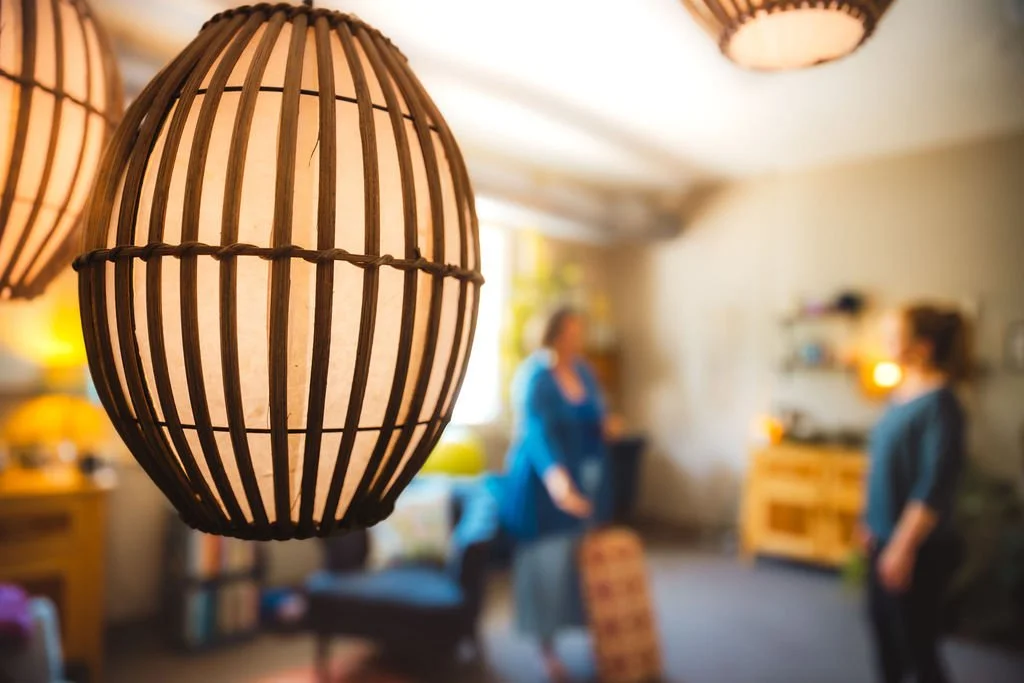A close-up of a hanging wooden lamp with a soft warm light in a cozy room, with two women talking in the background.
