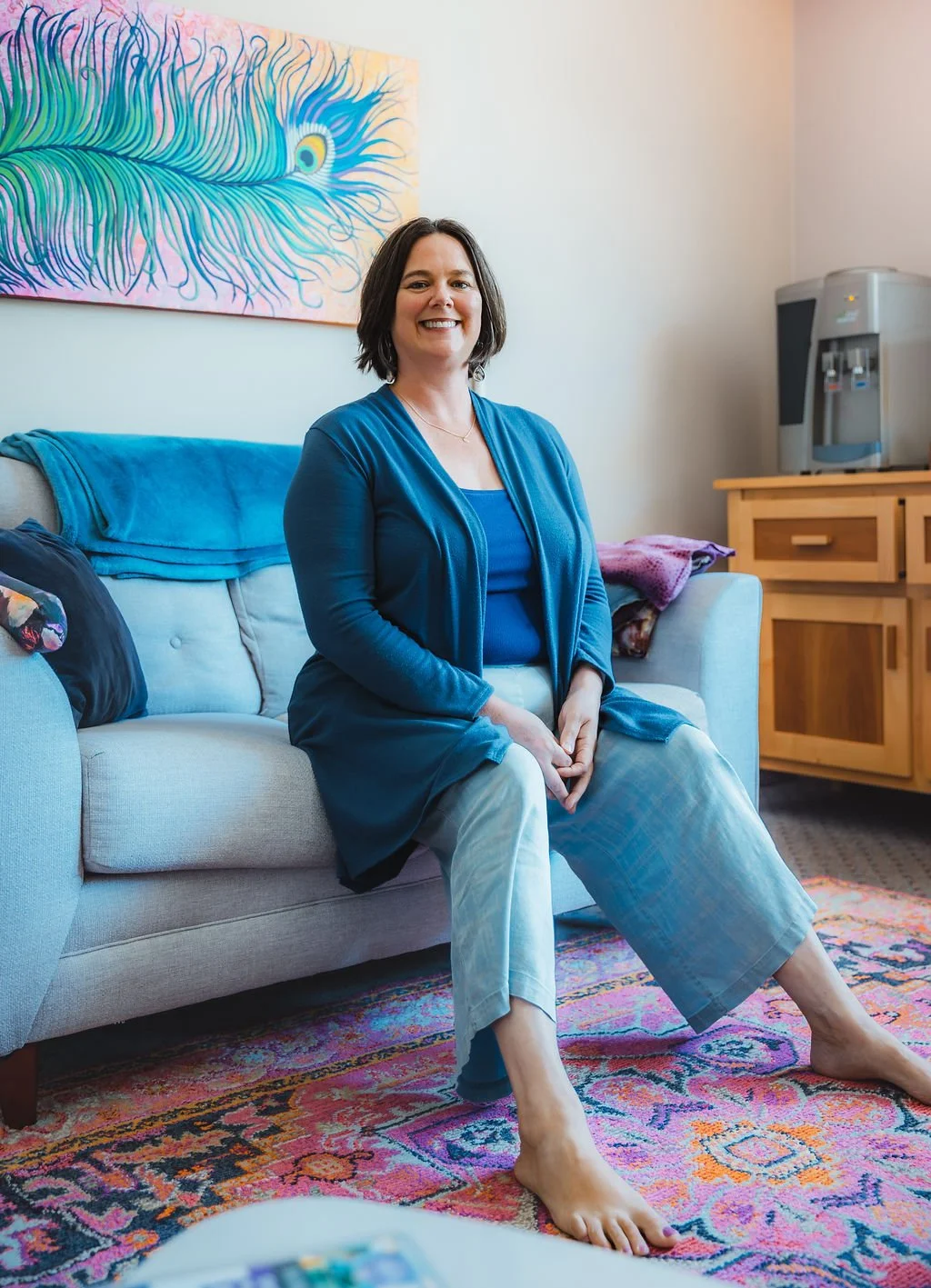 A woman seated on a light gray sofa in a living room, smiling at the camera. She is wearing a blue top and pants with a darker blue cardigan. The room has a colorful abstract feather painting on the wall, a carpet with vibrant patterns, and a wooden cabinet with a water cooler on top.