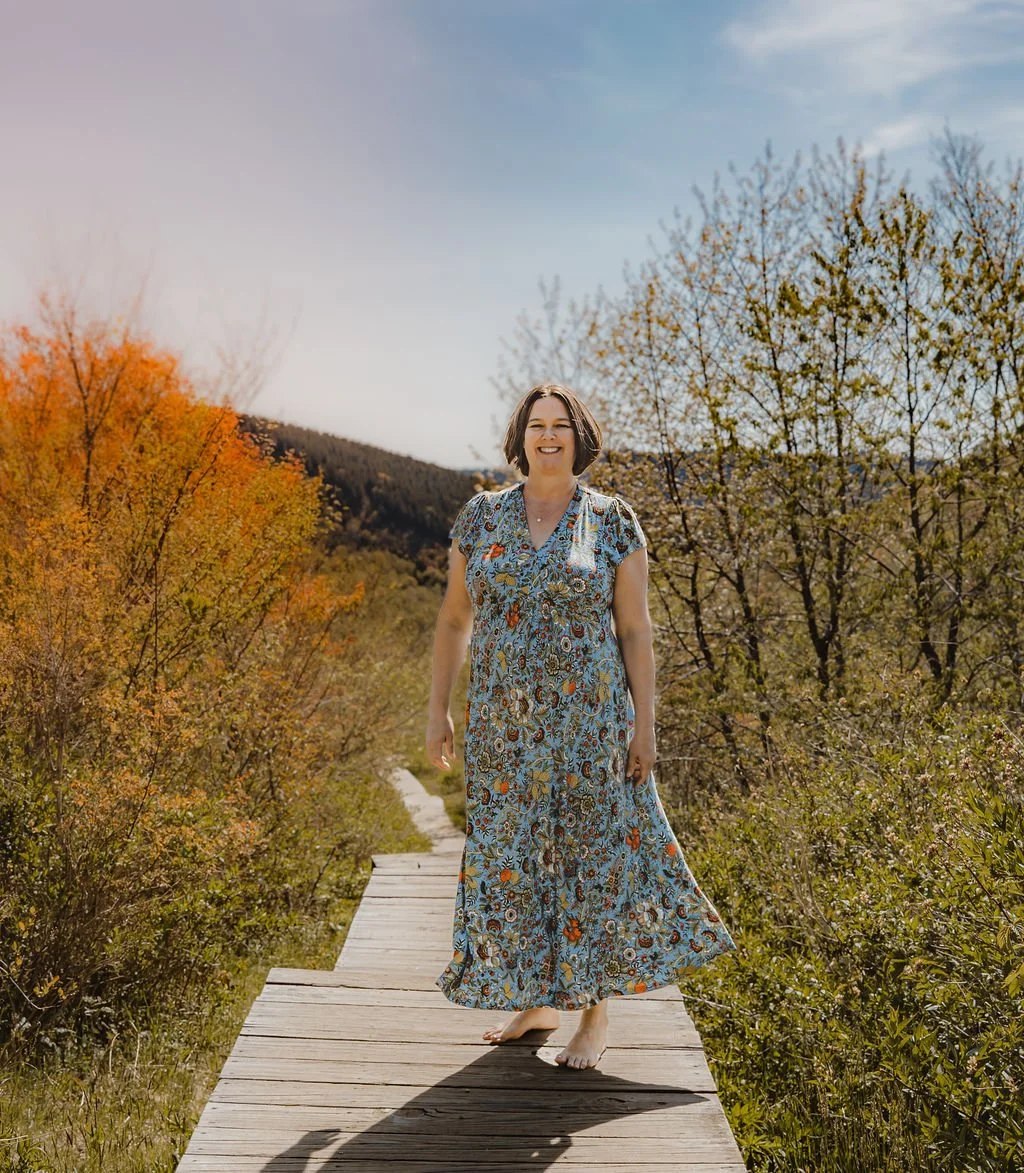 A woman smiling on a wooden walkway surrounded by trees with colorful fall foliage, wearing a long floral dress.