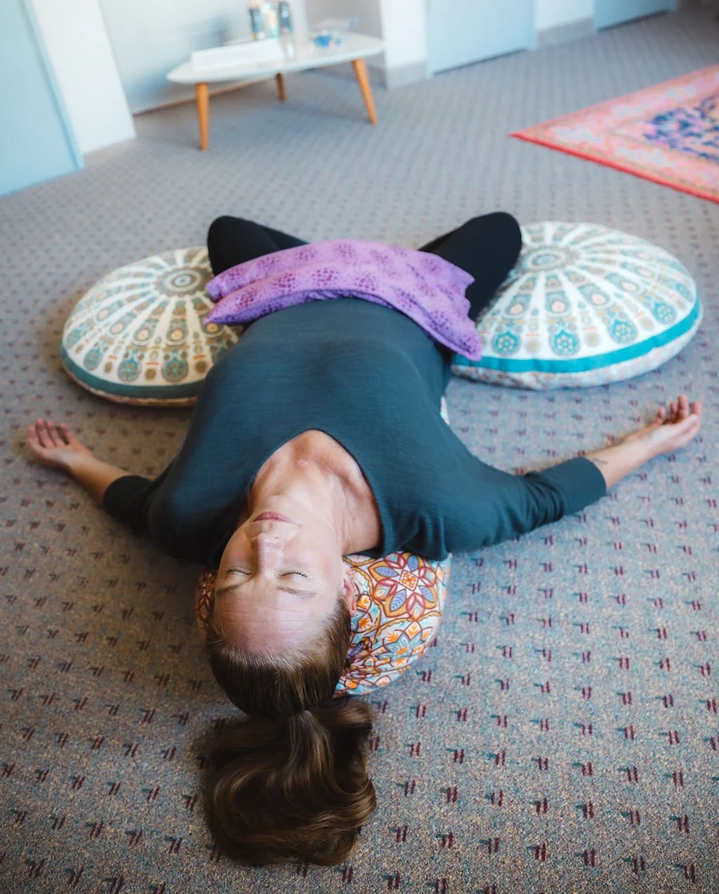 A woman lying on her back on the floor with eyes closed, head on a patterned round pillow, arms stretched out, with a purple blanket on her stomach, in a room with patterned carpet, a small table with bottles, and a colorful area rug.