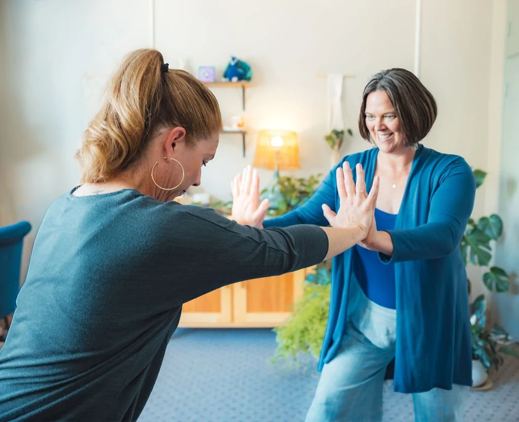 Heidi Andersen and coaching client, doing Chi for Two exercises, both smiling in a cozy, well-lit room with plants and decorative objects.