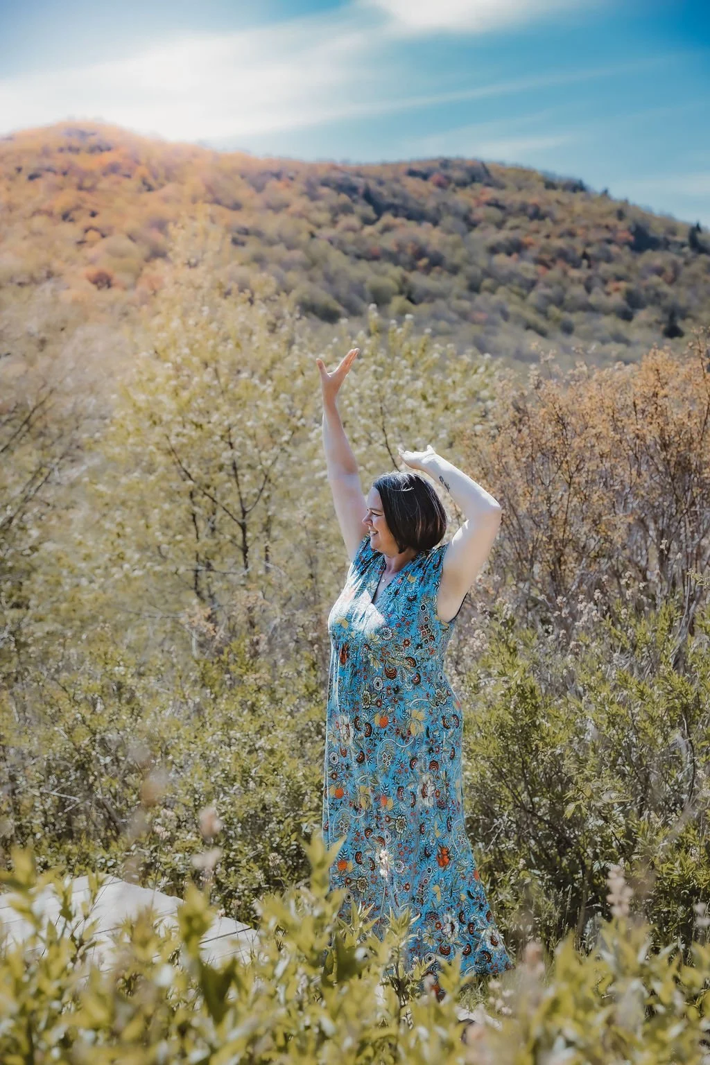 A woman in a blue floral dress smiling and raising her arms in a scenic outdoor setting with trees and mountains in autumn colors under a partly cloudy sky.