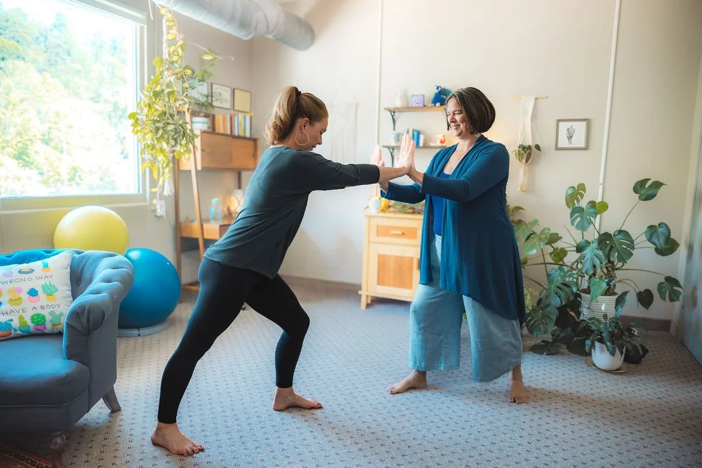 Two women practicing yoga or stretching together in a bright, cozy living room with plants and furniture.