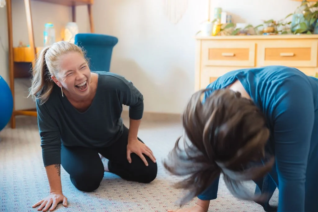 Two women laughing and playing on the floor, one woman kneeling and the other woman bending over, in a room with wooden furniture and plants.