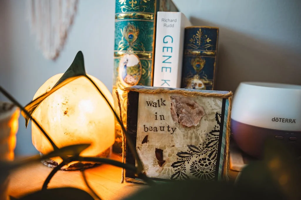 A lit Himalayan salt lamp, several books including "Gene K" by Richard Rudd and "Walk in Beauty" with decorative covers, a doTERRA essential oil diffuser, and a small decorative animal figure on a nightstand or shelf.