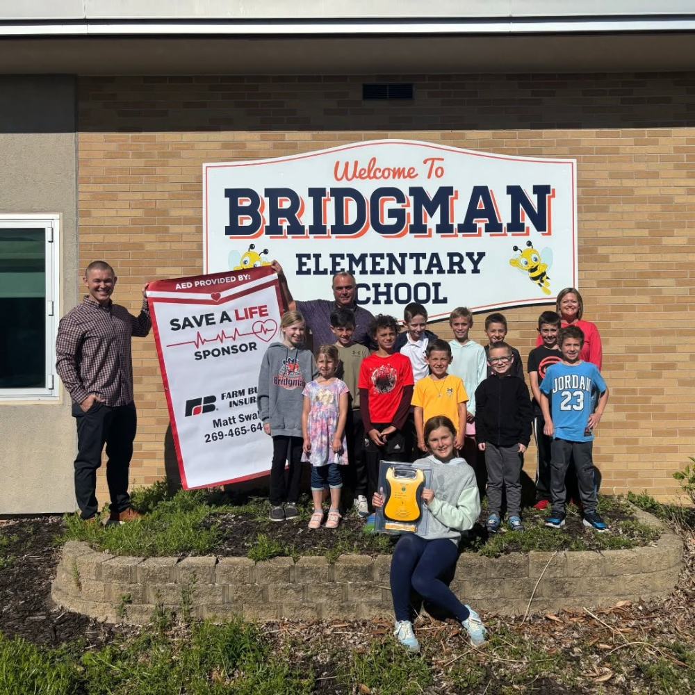 Group of children and adults standing in front of a school building with a large sign that reads 'Welcome to Bridgman Elementary School'. A woman in front is holding a yellow device and there's a person on the left holding a sign about an AED sponsor.