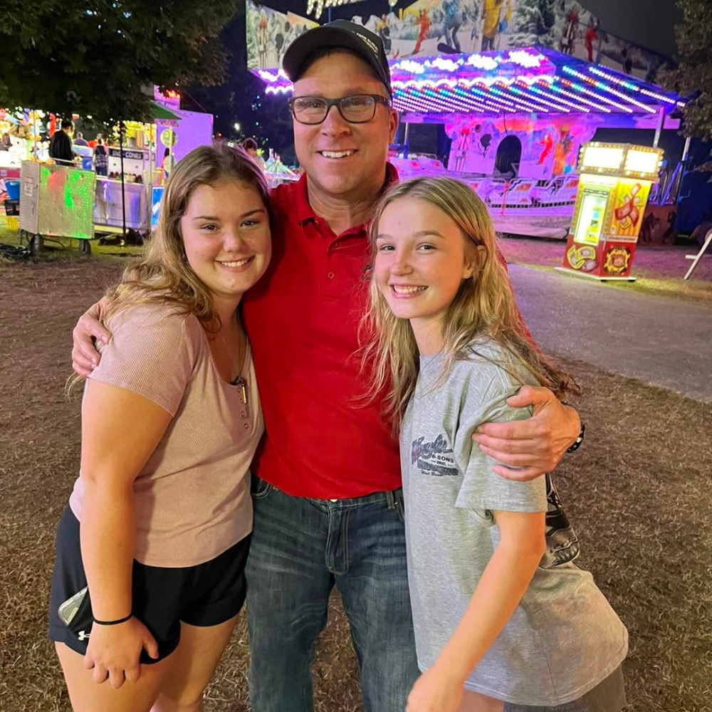 Two girls and a man smiling at a carnival at night with the man and girls hugging and the carnival rides and lights in the background.