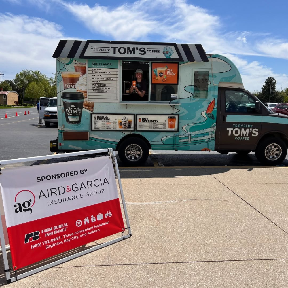 A teal and white coffee food truck named 'Travelin' Tom's Coffee' is parked on a street. The truck advertises various coffee drinks and frozen treats. There is a person inside the truck serving coffee. A sign in the foreground promotes insurance coverage sponsored by Aird & Garcia Insurance Group.