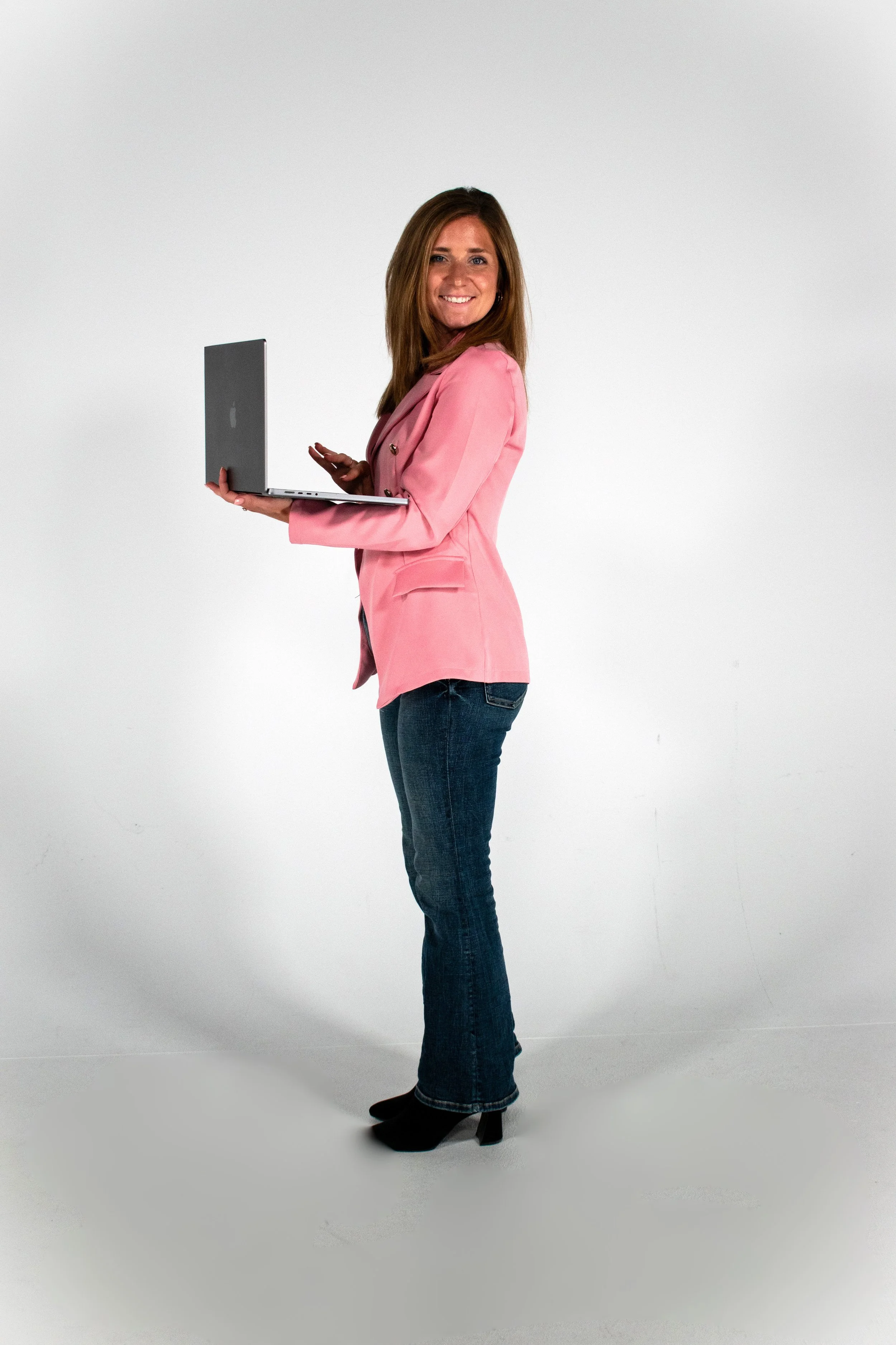 A woman standing sideways, holding a laptop, wearing a pink blazer, blue jeans, and black heels, smiling at the camera, against a plain white background.