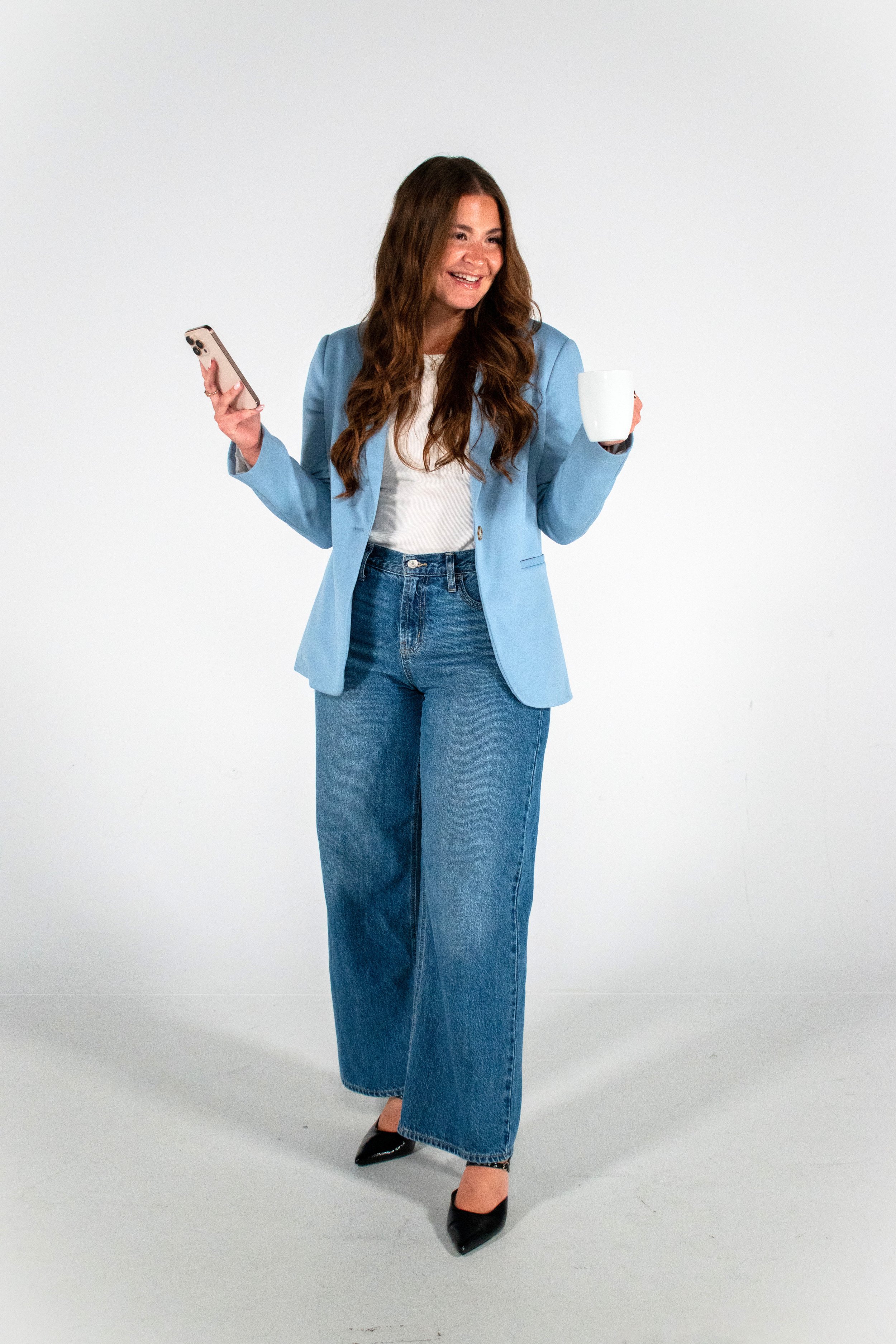 A woman in a light blue blazer, white top, and wide jeans holding a phone in one hand and a white mug in the other, smiling against a plain white background.