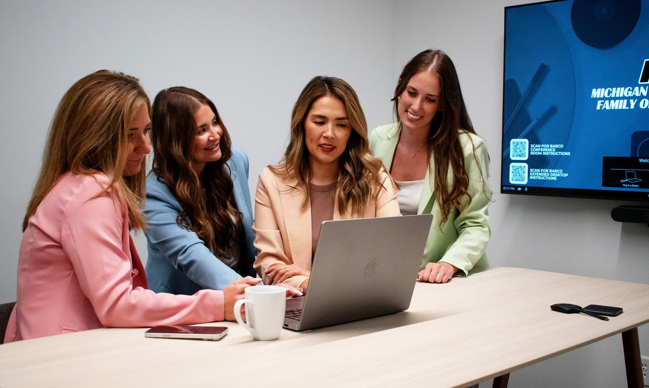Five women in business attire gathered around a table looking at a laptop, with a large digital screen on the wall showing 'Michigan Family'.
