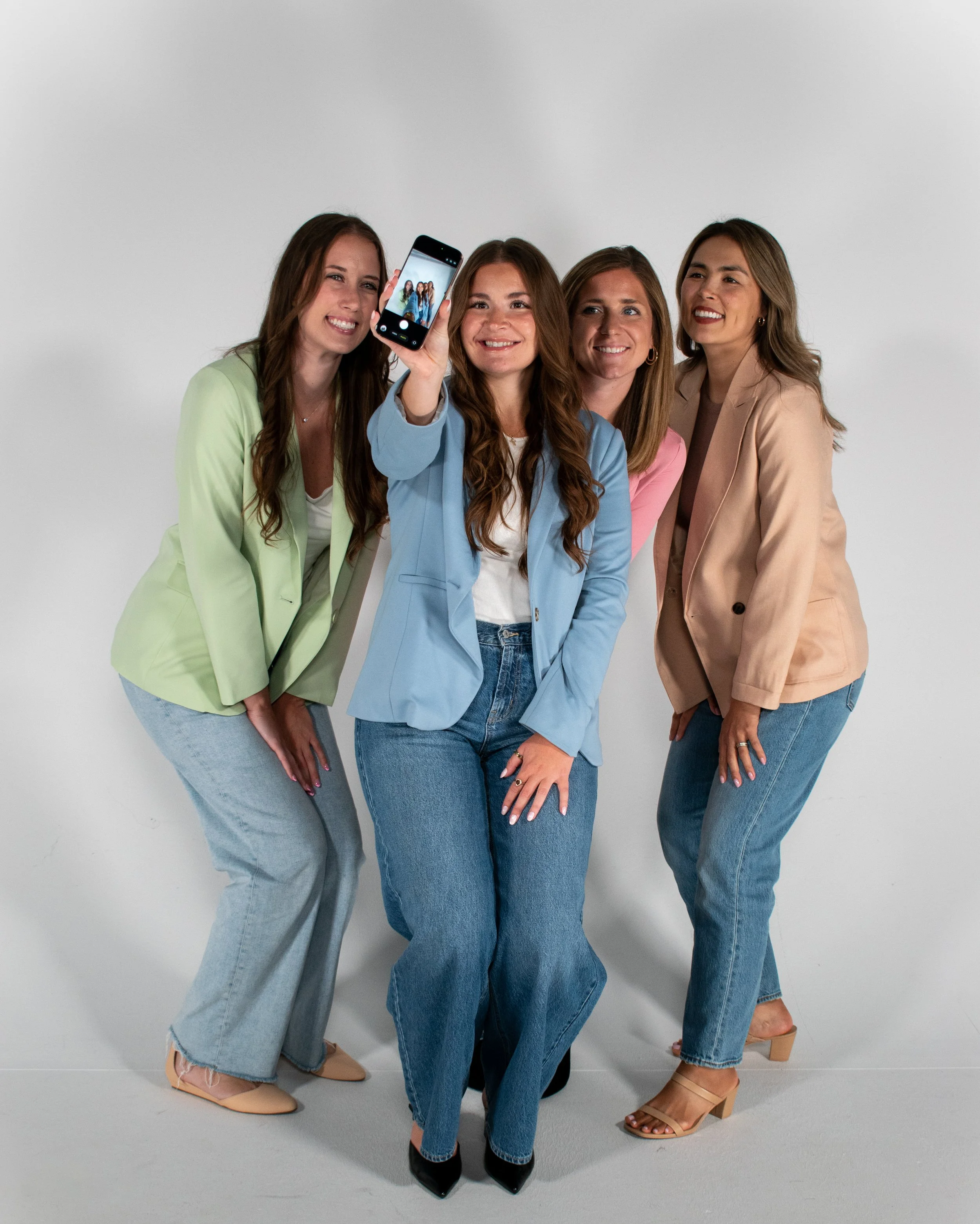 Four women taking a group selfie against a plain white background, all smiling and dressed in pastel-colored blazers and blue jeans.