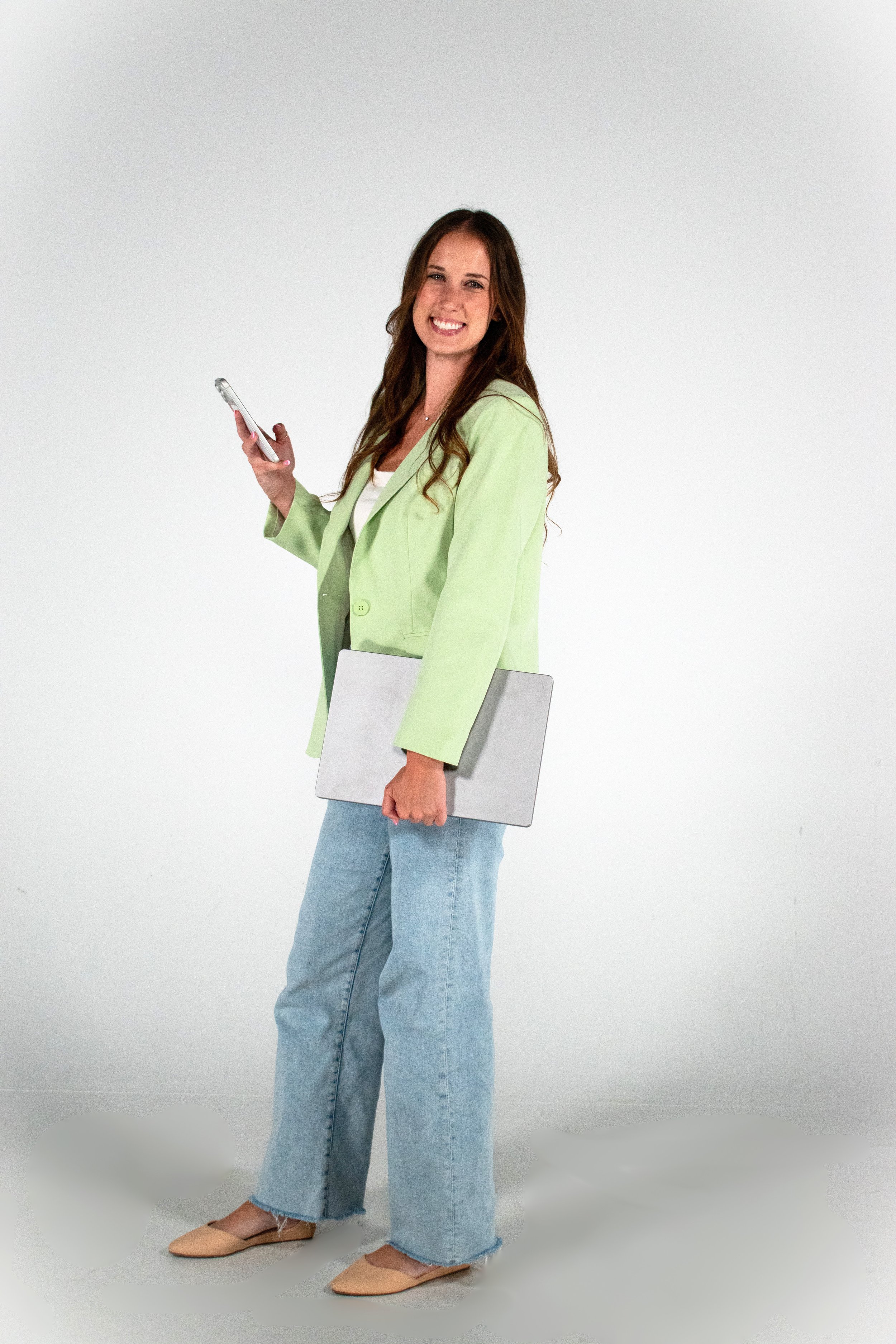 A woman standing against a plain white background, smiling, holding a smartphone in one hand and a silver laptop under her other arm. She is wearing a light green blazer, a white top, blue jeans, and beige flats.