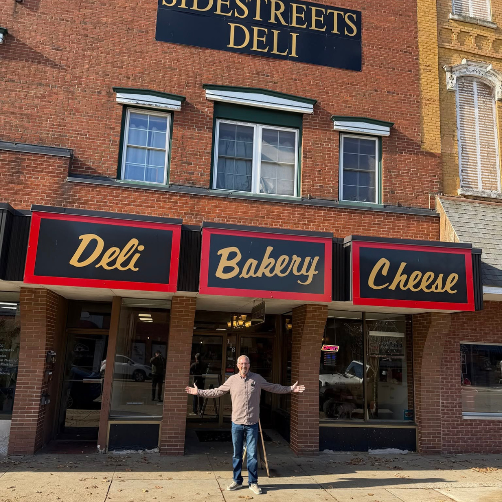 A man standing outside a deli, bakery, and cheese shop with a sign that reads 'Deli Bakery Cheese'. The storefront has brick walls and three signs in black with gold lettering. The man is smiling, wearing glasses, and gesturing with his arms open.