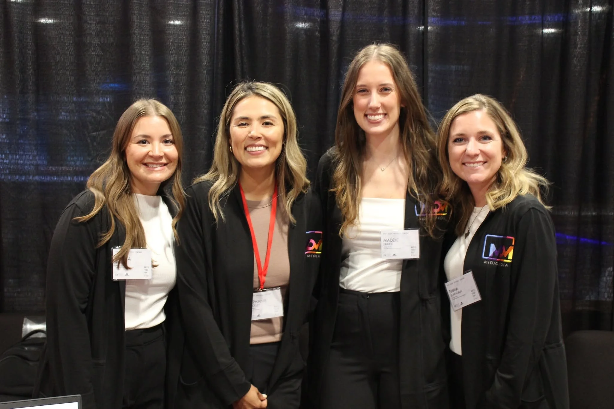 Four women standing together at an event, smiling for the photo. They are wearing black jackets with a colorful logo and name tags.