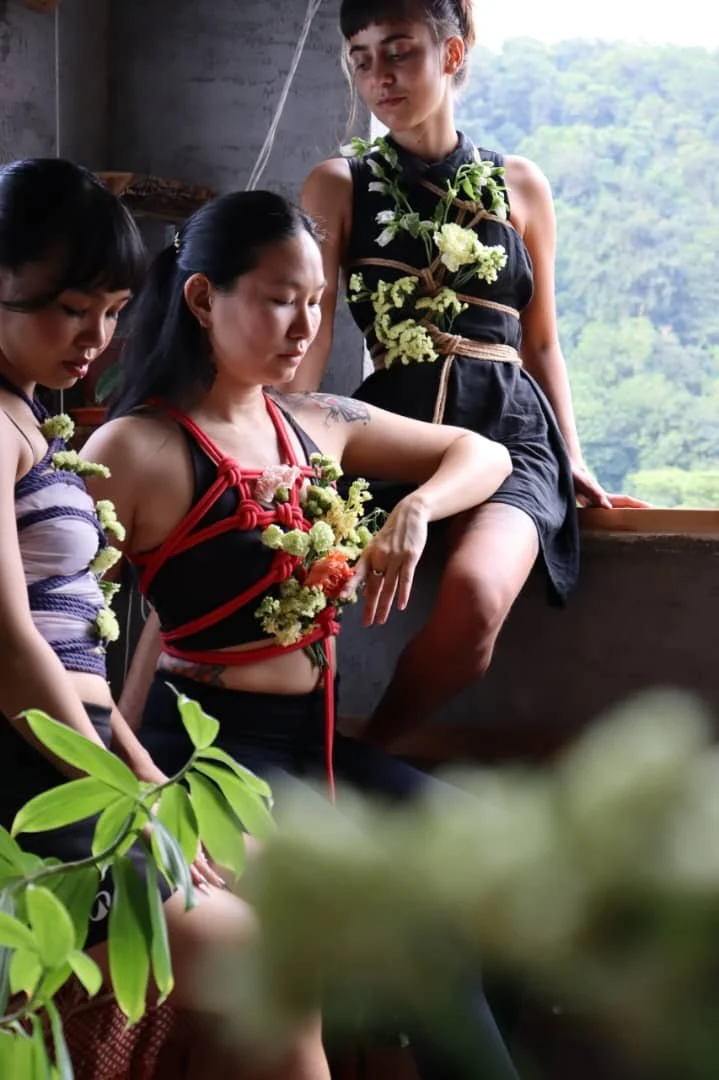 Three women, two young and one older, are in a relaxed setting, surrounded by greenery outside a rustic structure. The woman in the center is wearing a black top with red rope, adorned with flowers, and appears contemplative. The other two women are dressed in traditional attire with flowers, and they are observing or participating in a cultural or spiritual activity.