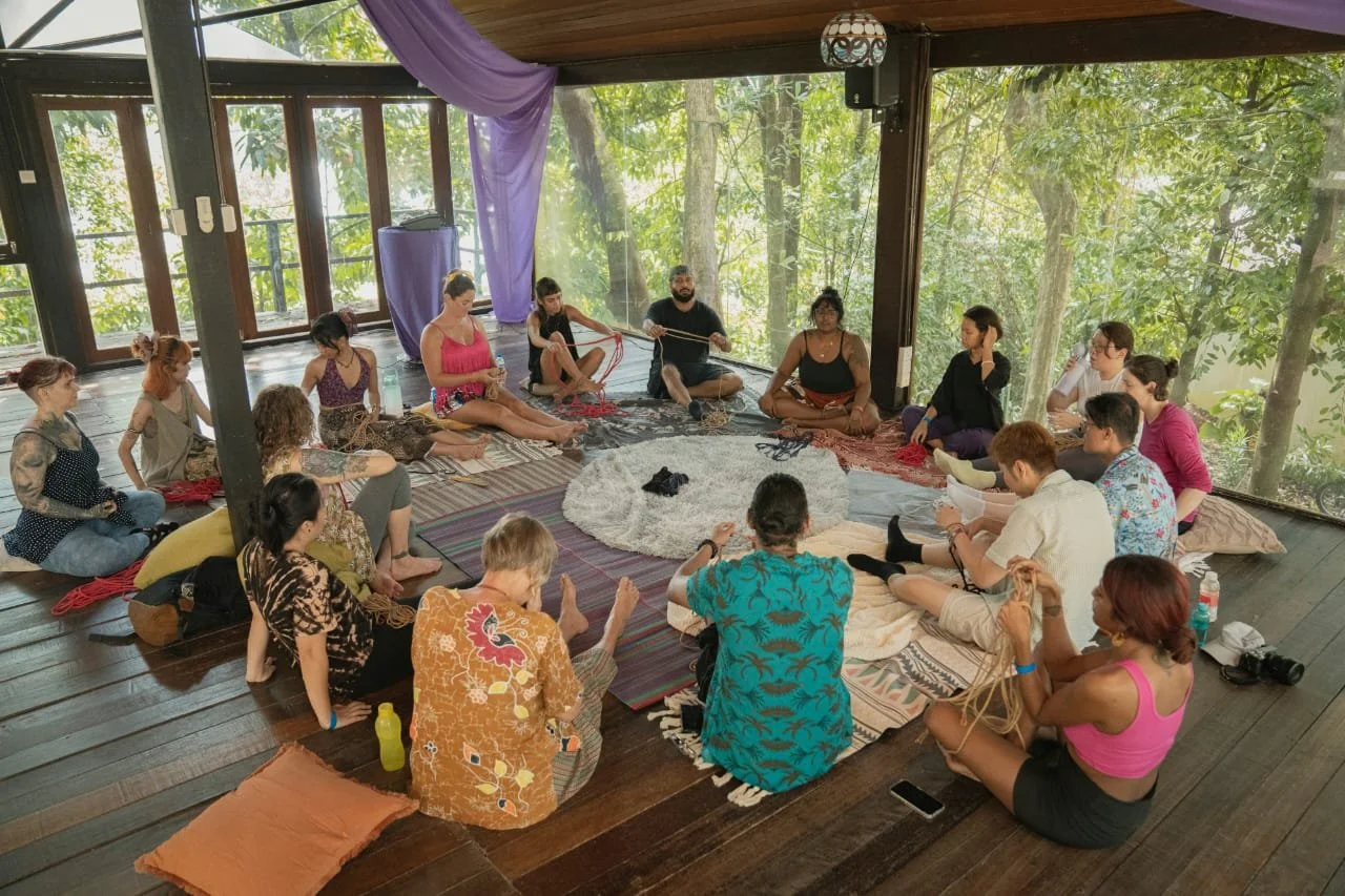 Group of people sitting in a circle on wooden floor inside a room with large windows overlooking trees, participating in a self tying shibari workshop in bumi fest