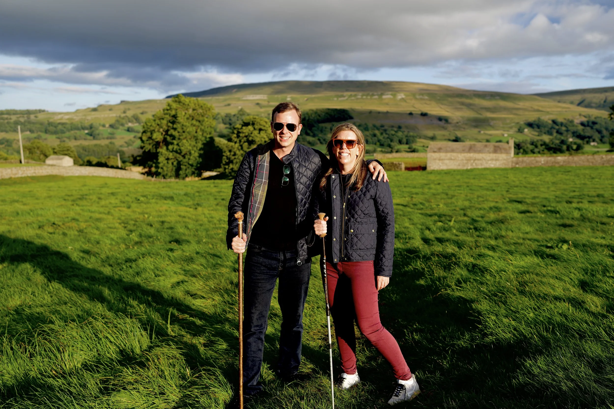 A man (Steve Johnson) and a woman (Brooke Van Dam) standing with walking sticks in green field with rolling hills on a cloudy day.
