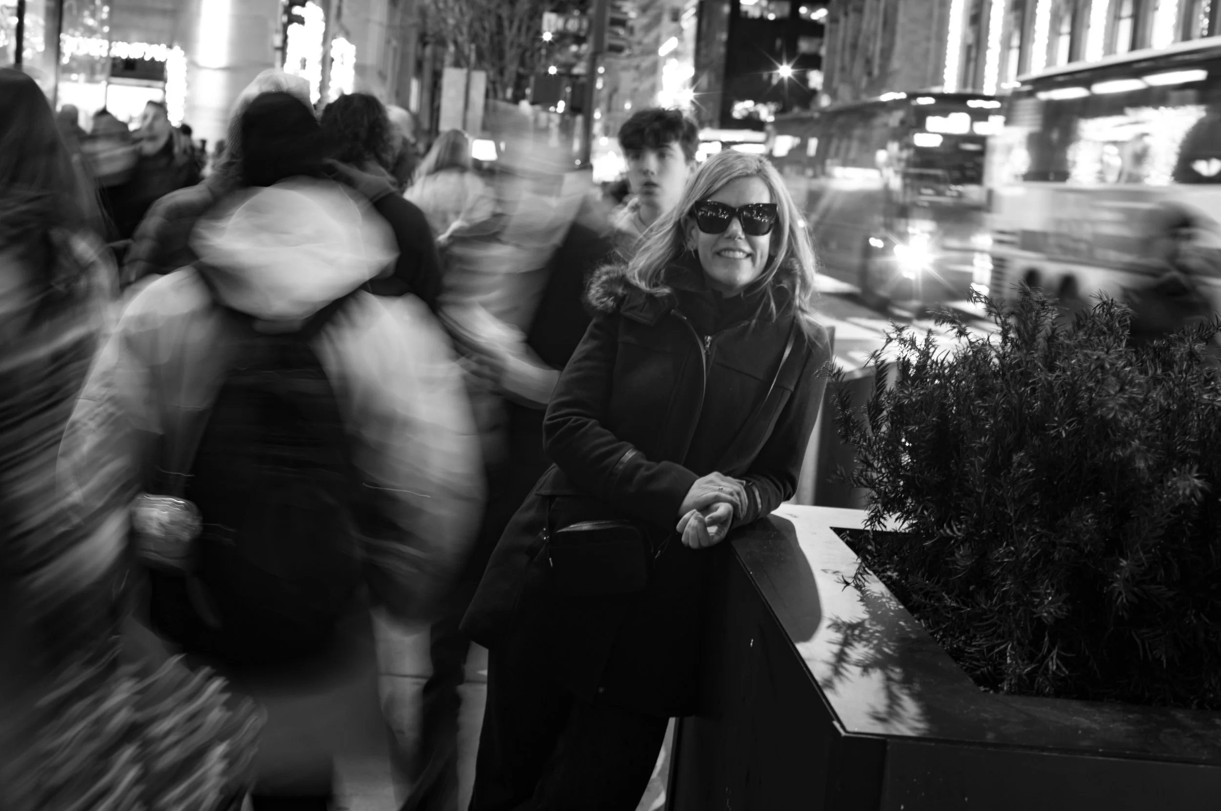 A black and white photo of a blonde woman, Brooke Van Dam, standing on the streets of New York City with blurry people, cars and busses going by.