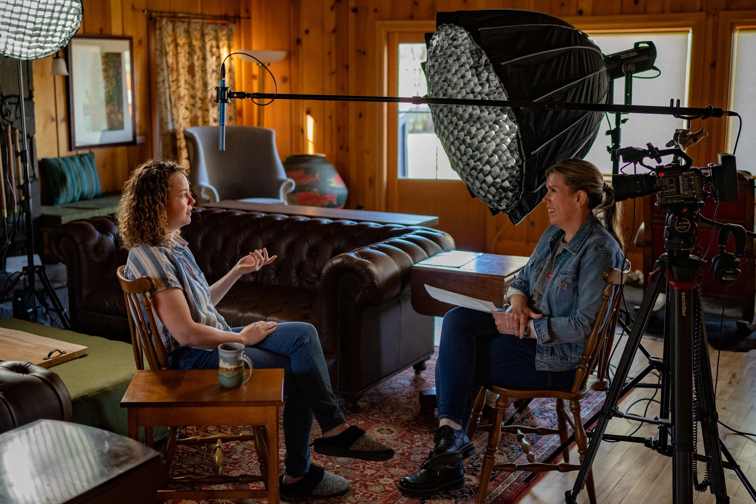 Two women sitting down, one being interviewed with lights and cameras.