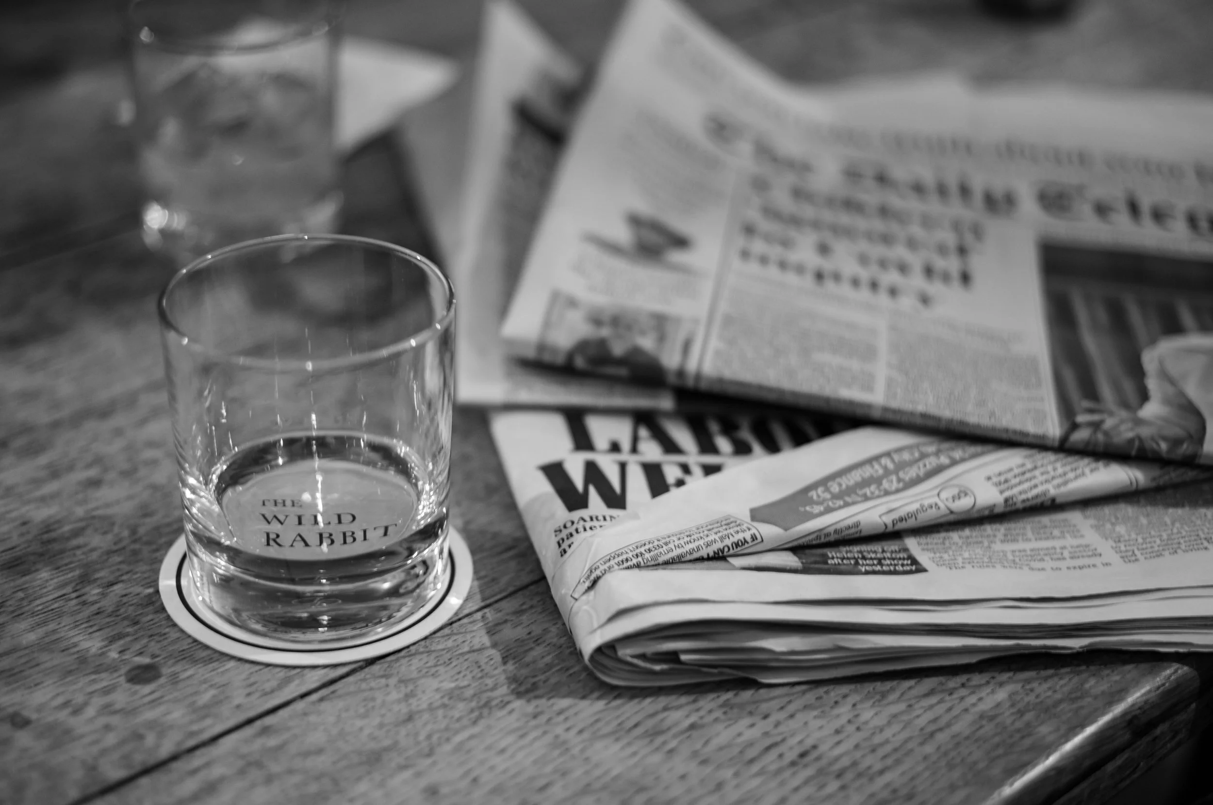 A black and white photo of a stack of newspapers with a glass empty sitting next to it on an old wooden table.