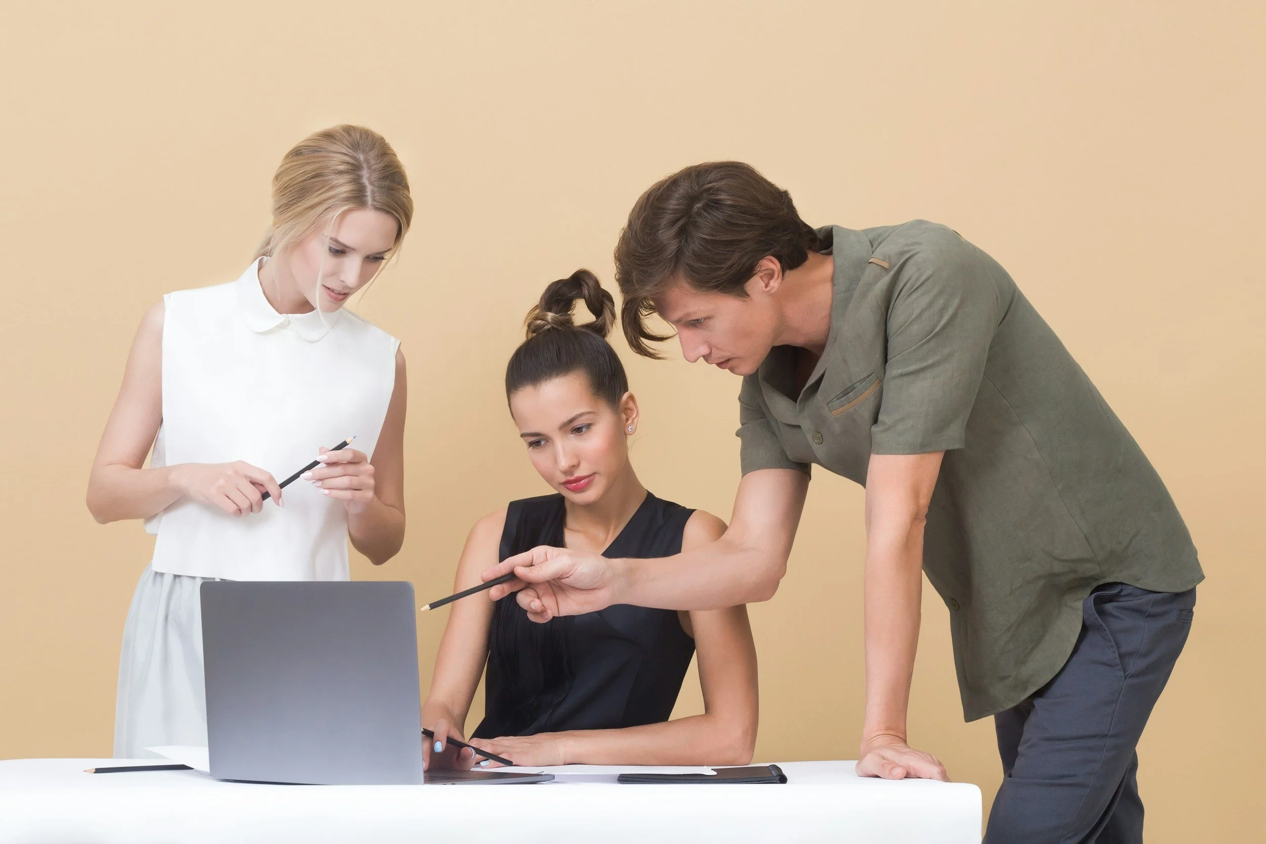 Four people are gathered around a laptop, discussing something, with two women standing and one man leaning over the table, on a beige background.