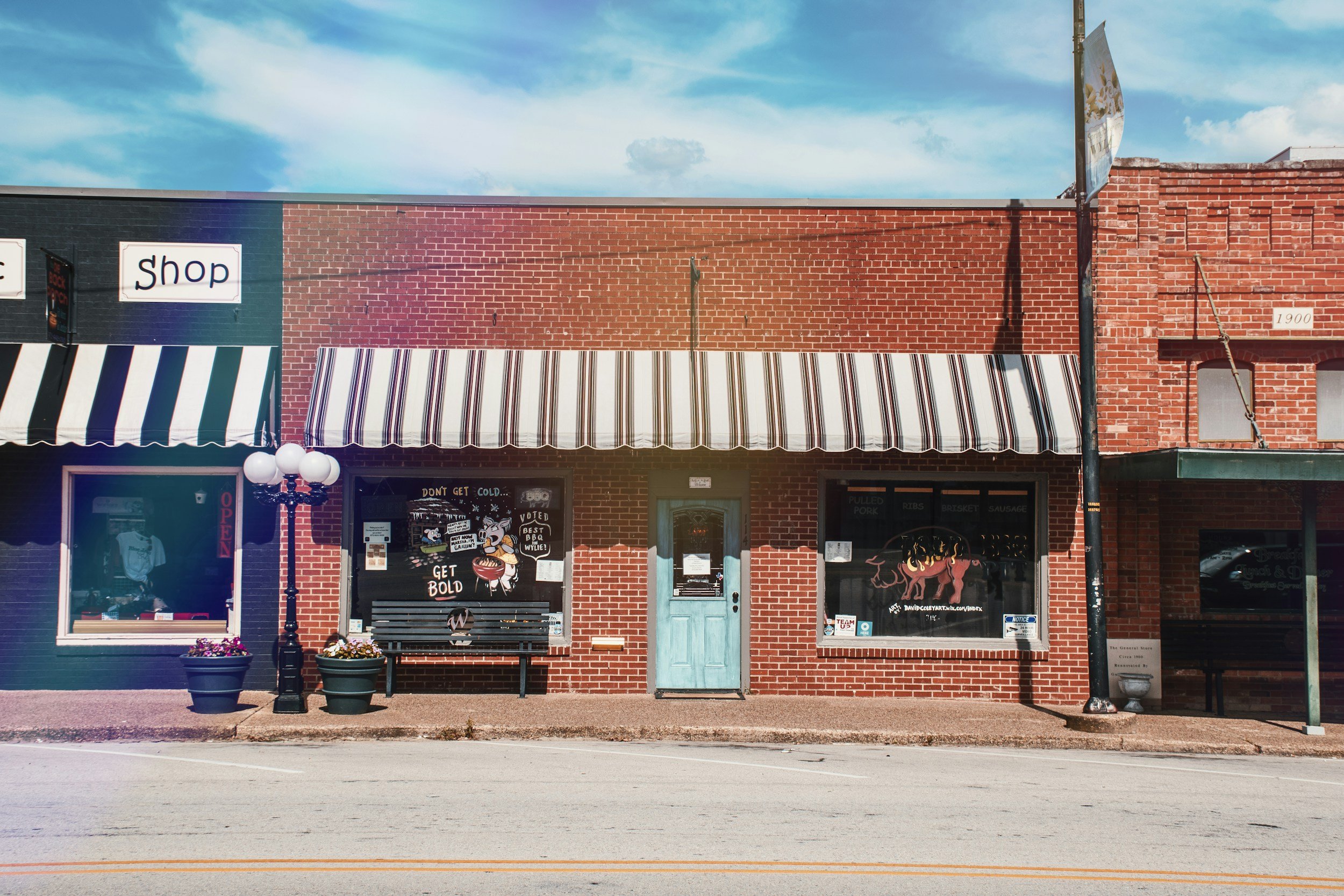 The storefront of a brick building with a black and white striped awning. There are two large windows, a turquoise door, and a sign that reads "Shop." A black lamppost with white globe lights is in front of the building, along with two large flower pots, a bench, and a sidewalk. The sky is blue with some clouds.