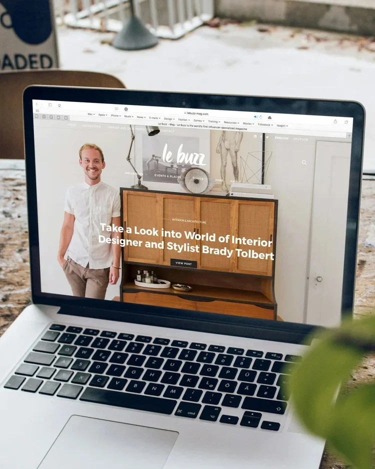 A silver MacBook laptop displaying a webpage about interior design and an interior designer named Brady Tolbert. The screen shows a smiling man in a white shirt standing next to a wooden cabinet. The laptop is placed on a wooden table with a snowy outdoor background and some blurred green leaves in the foreground.
