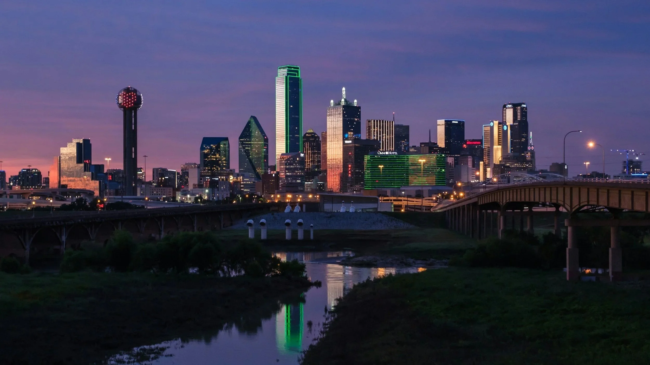 Nighttime view of Dallas downtown skyline with illuminated buildings, a river reflecting city lights, and a bridge in the foreground.