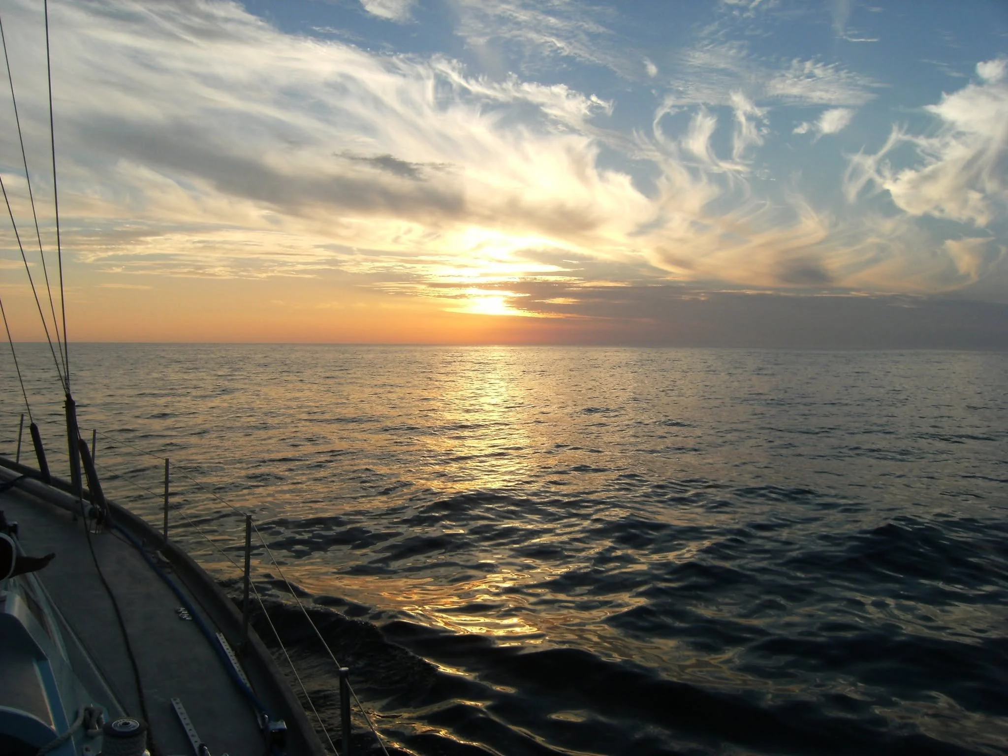 Sunset over the ocean viewed from a boat with part of the boat's rigging in the foreground and wispy clouds in the sky.