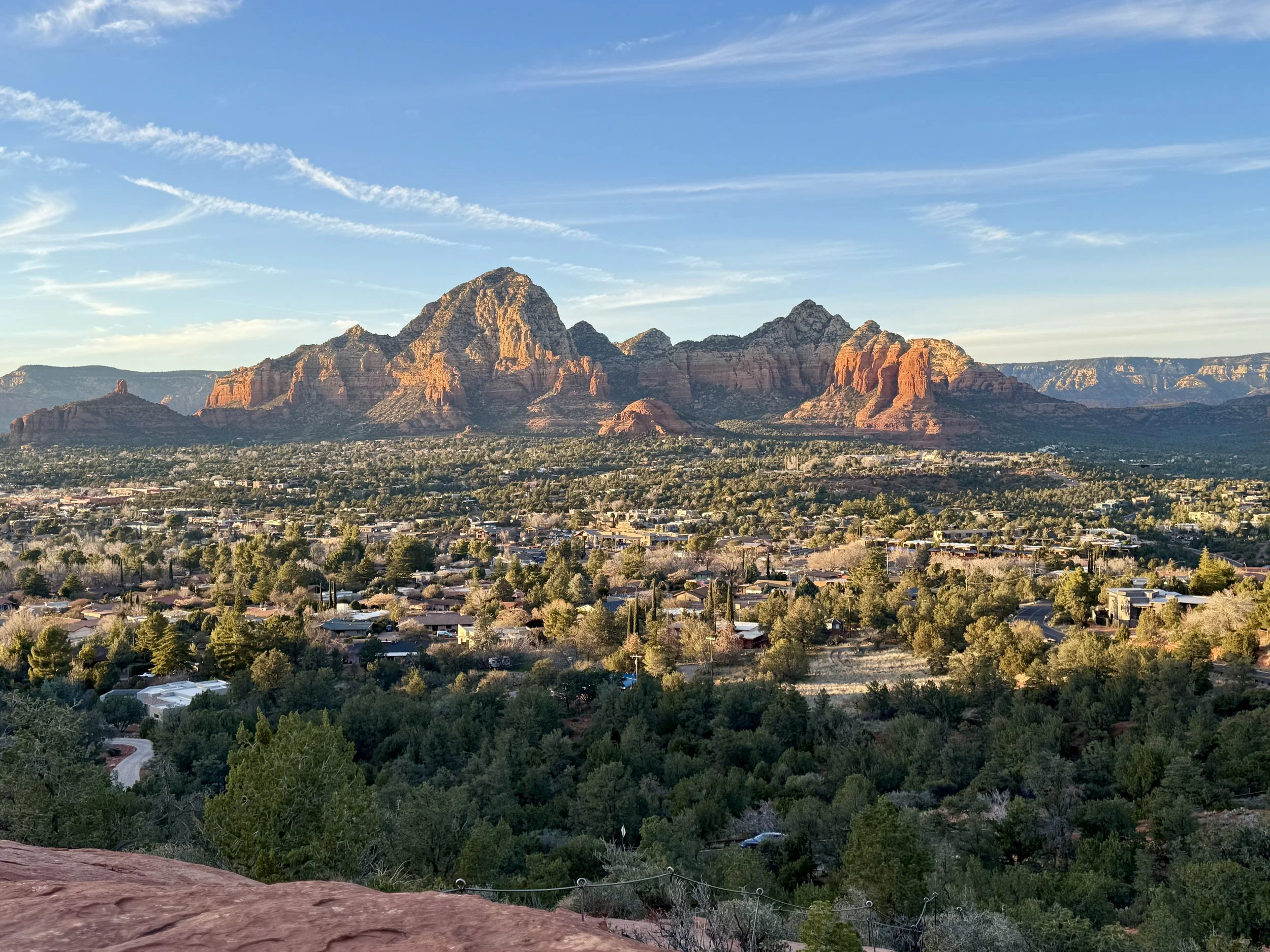 A panaromic view of Sedona, Arizona at golden hour looking east from Airport Mesa.  In the distance are Twin Buttes, the red rock landscapes, and wispy white clouds against the light blue backdrop of the sky