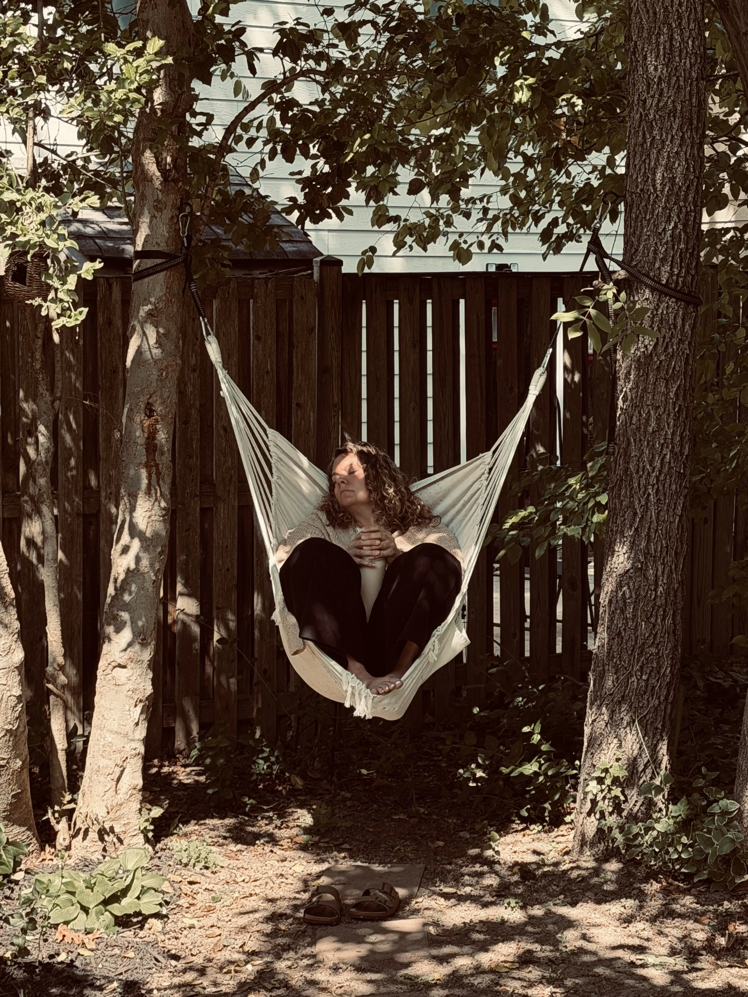 Person with curly hair sitting in a hammock hanging between two trees in a backyard, with a wooden fence and plants in the background. Shoes are on the ground beneath the hammock.