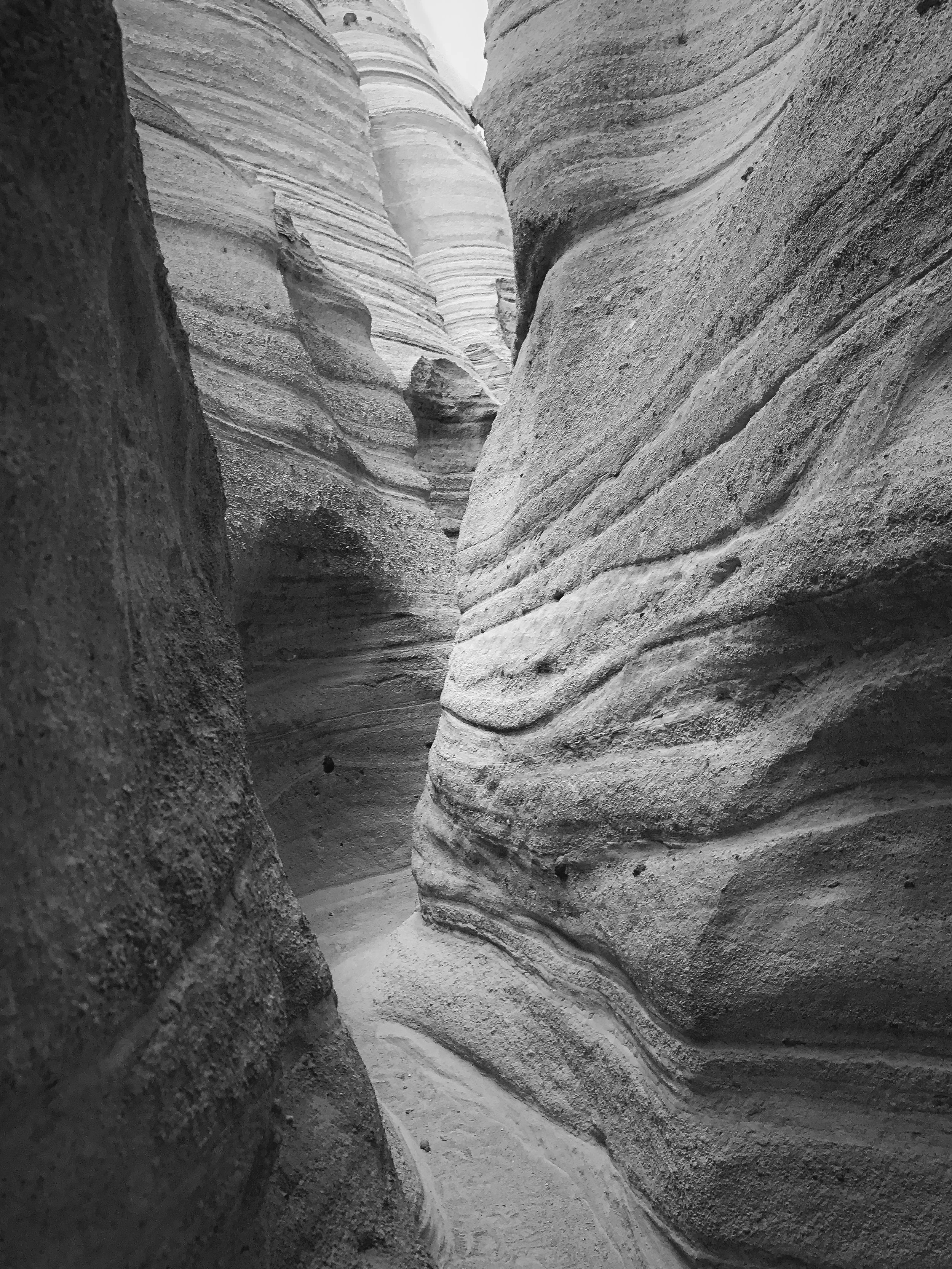 Black and white photo of narrow canyon walls with layered, textured rock formations, showing a winding trail through the slot canyon at Kasha-Kutuwe Tent Rocks in New Mexico.