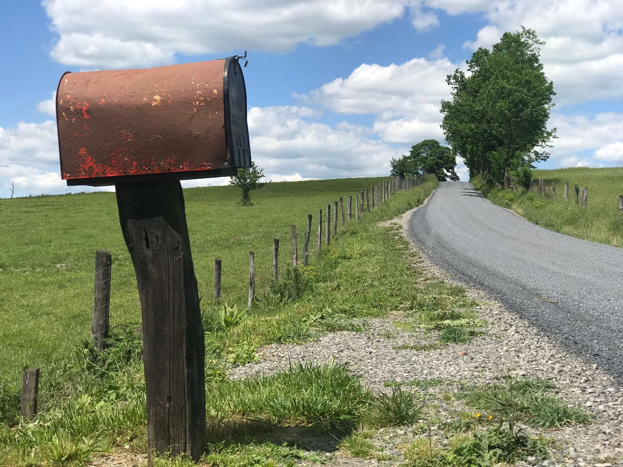 A rural gravel road lined with a wire fence on the left side, leading towards a large green tree and a blue sky with white clouds. On the left side, there's an old, rusted mailbox on a wooden post. In Highland County, Virginia.