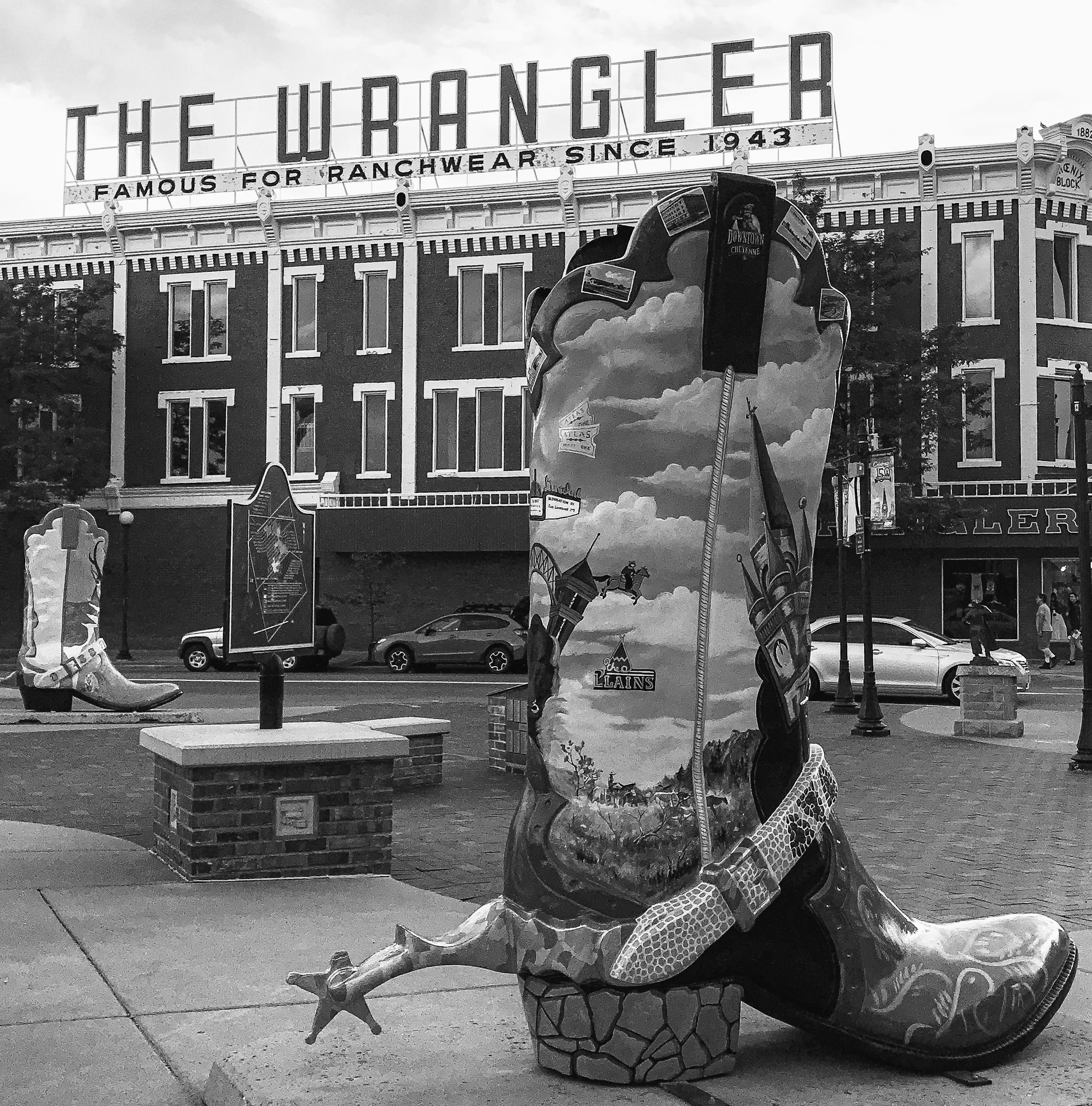 Black and white photo of The Wrangler Store on Capitol Avenue in Cheyenne, Wyoming featuring two of the cowboy boot  sculptures painted by local artists. Cars are parked along the street in front of the store.