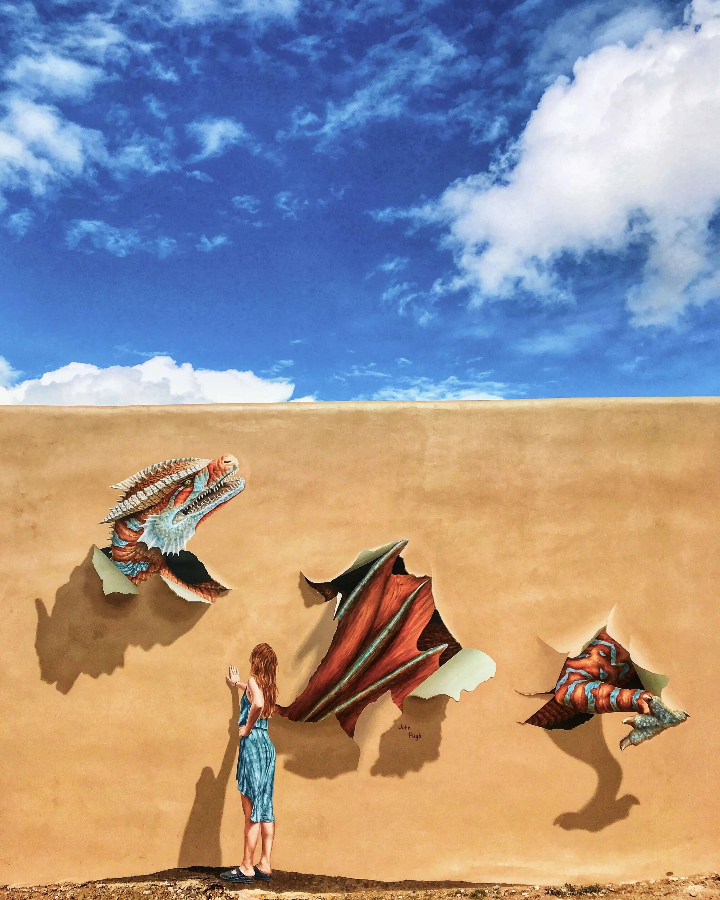 Color photograph with deep blue skies and white clouds over an adobe wall of the Jean Cocteau theater in Santa Fe, NM featuring a John Pugh mural of a red-headed young girl looking up at a dragon that is painted as if breaking through the wall