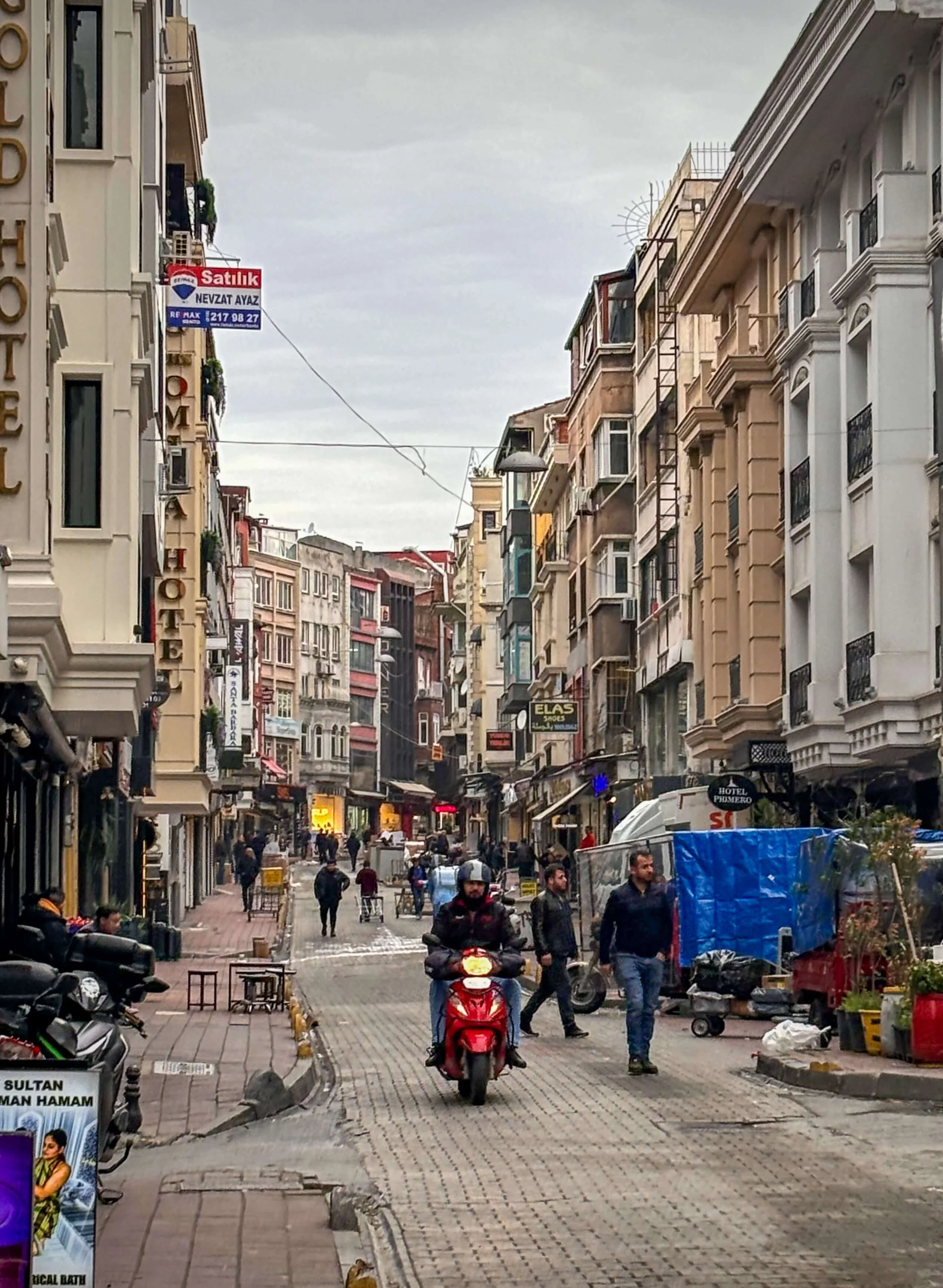 Morning streets in the old city section of Istanbul, Turkey with pedestrians walking cobbled streets and a rider on a moped passing by shops, a hotel, and other packed together buildings under a gray sky