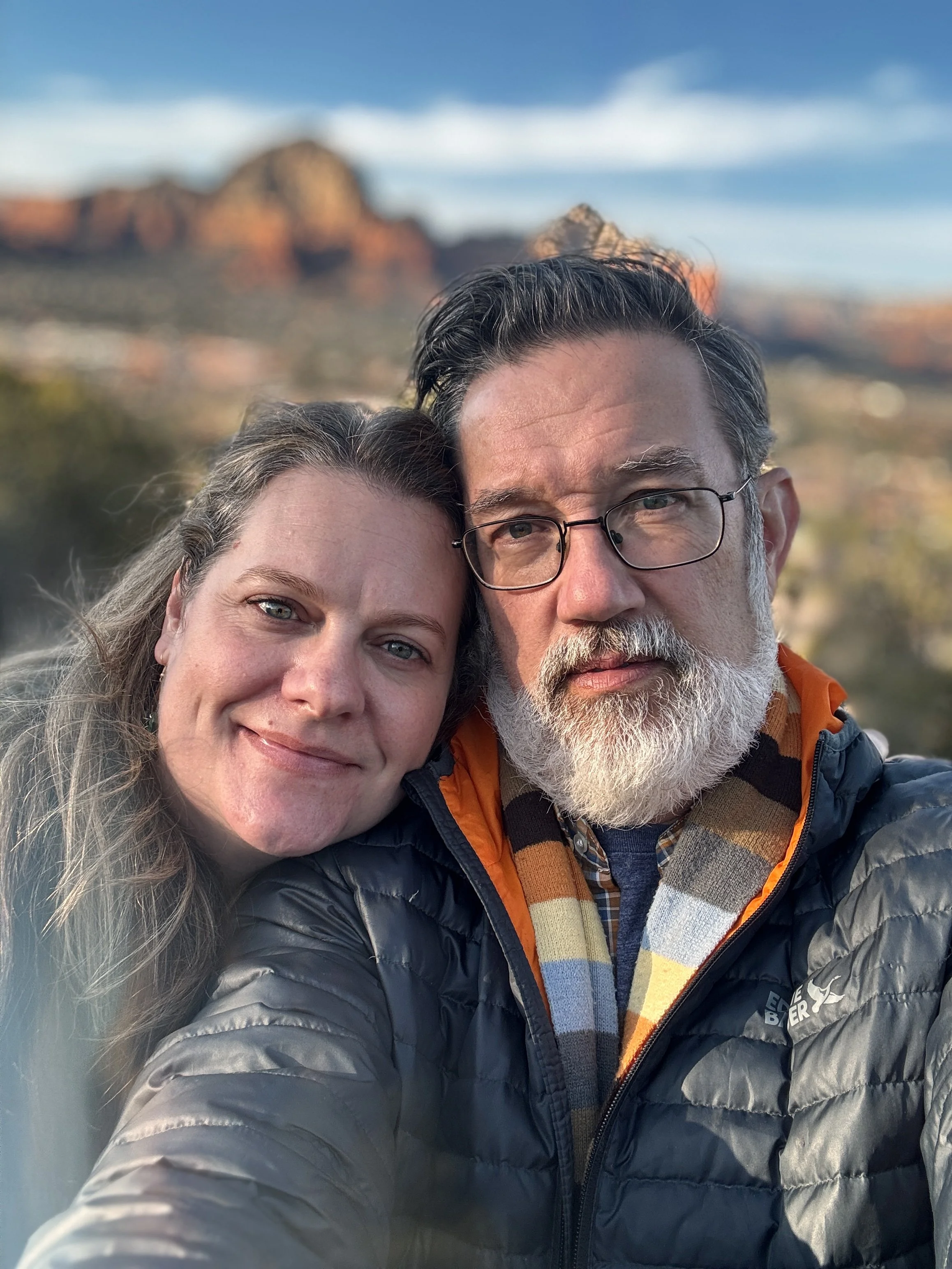 A close-up photo of a smiling woman and a serious man with glasses, both wearing winter jackets, outdoors with blurred mountain scenery and blue sky in the background.