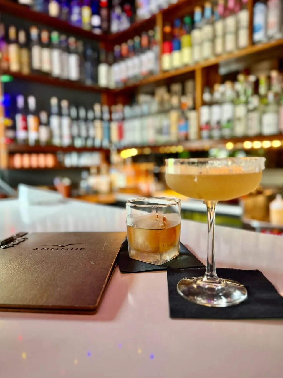 A mezcal old fashioned and a craft cocktail served in a rimmed coupe on the bar at Anoche in Asheville, NC, bottle-lined back bar in soft focus