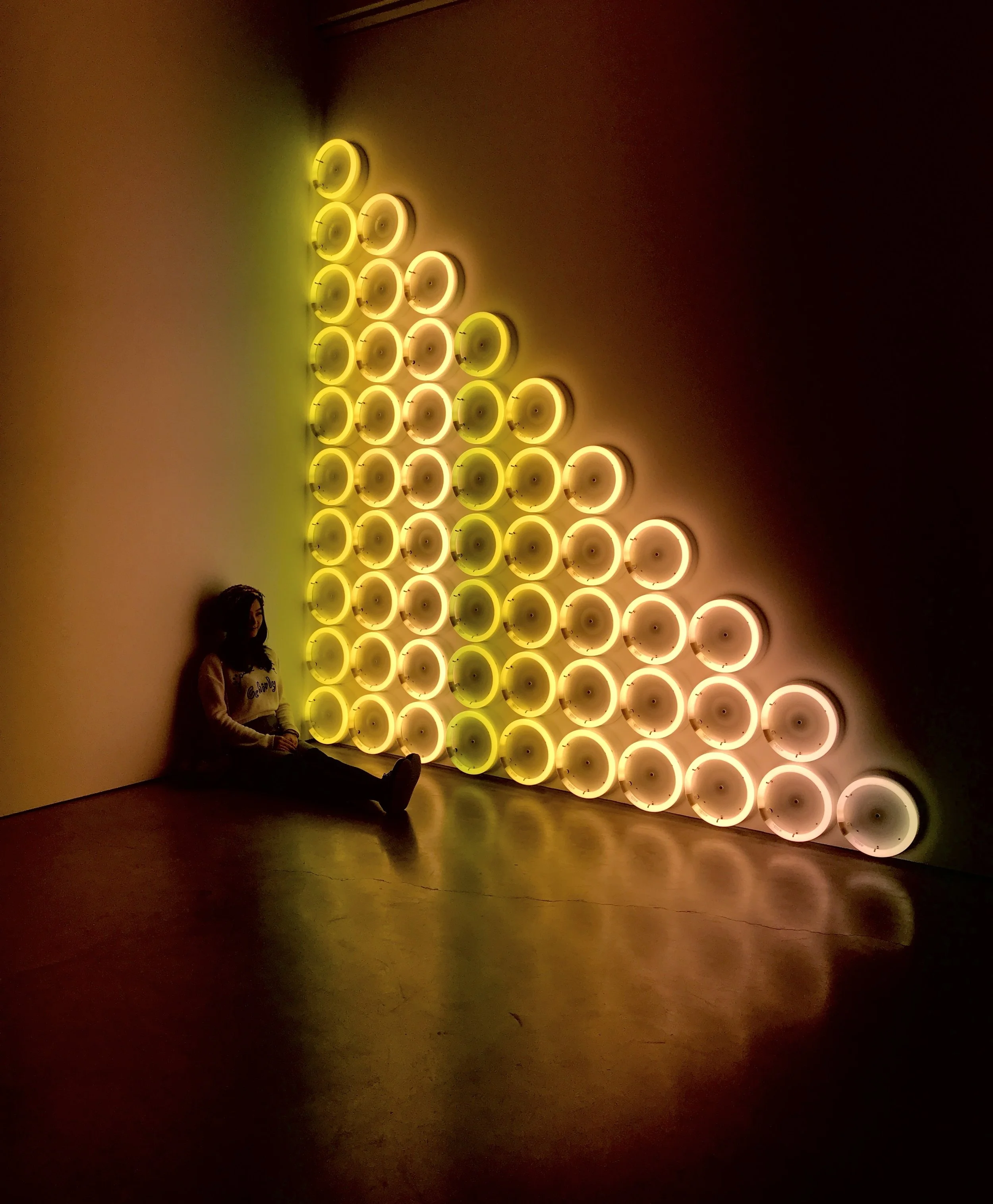 Woman sitting against a wall beside a triangular installation of glowing yellow and white circular lights