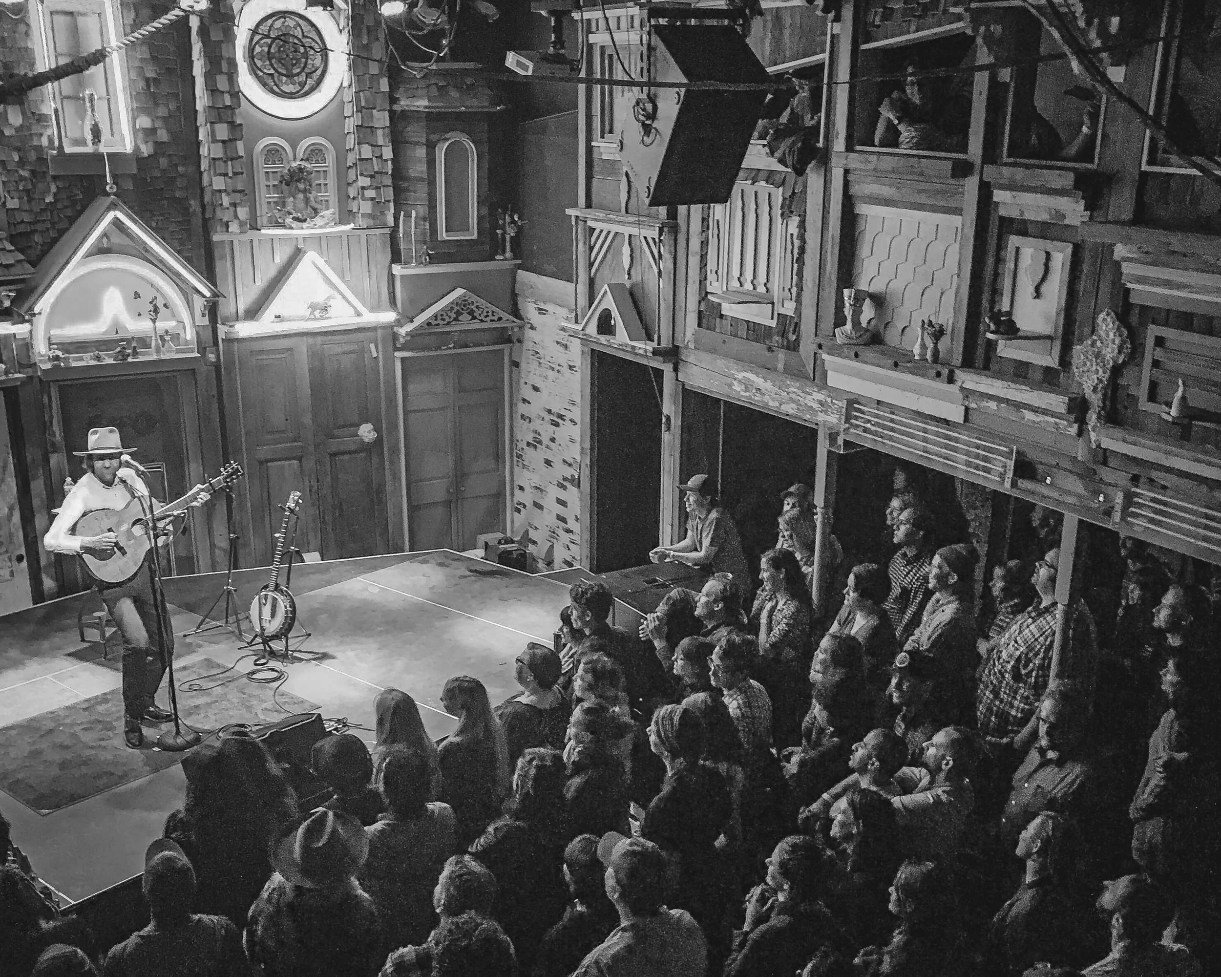 A black and white photograph of folk musician Willie Watson performing with a guitar in hand on the stage at Meow Wolf in Santa Fe, NM as a crowd of onlookers watches from below and to the right of the stage.