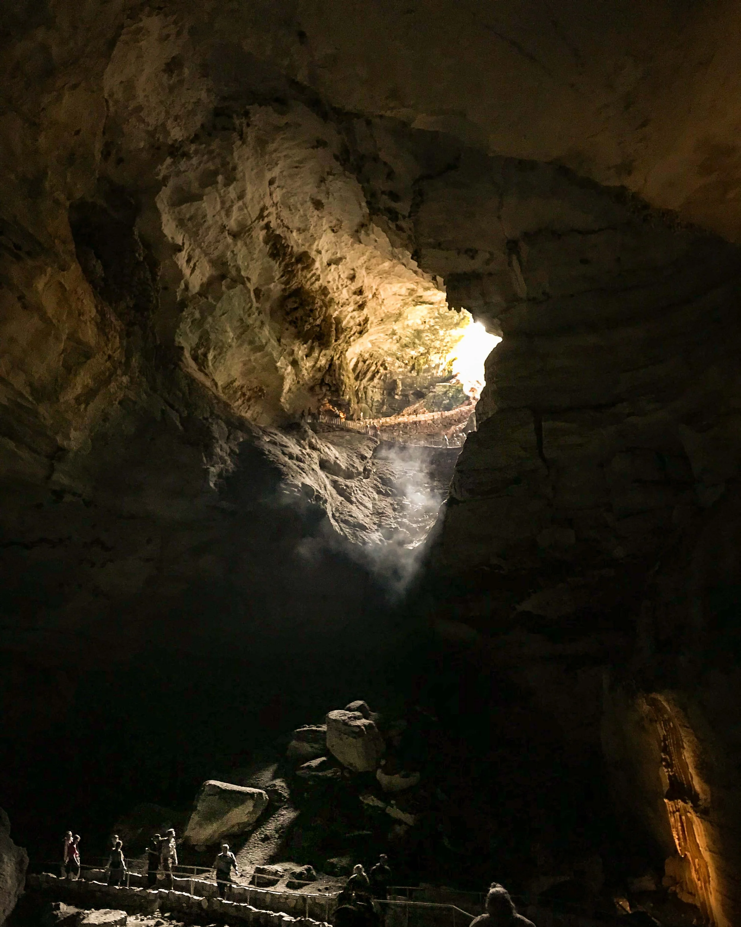 Color photograph of the mouth of Carlsbad Cavern National Park in New Mexico with golden sunlight streaming in through onto rock walls while below the light are park visitors looking small by comparison in the darker lower portion of the photograph.