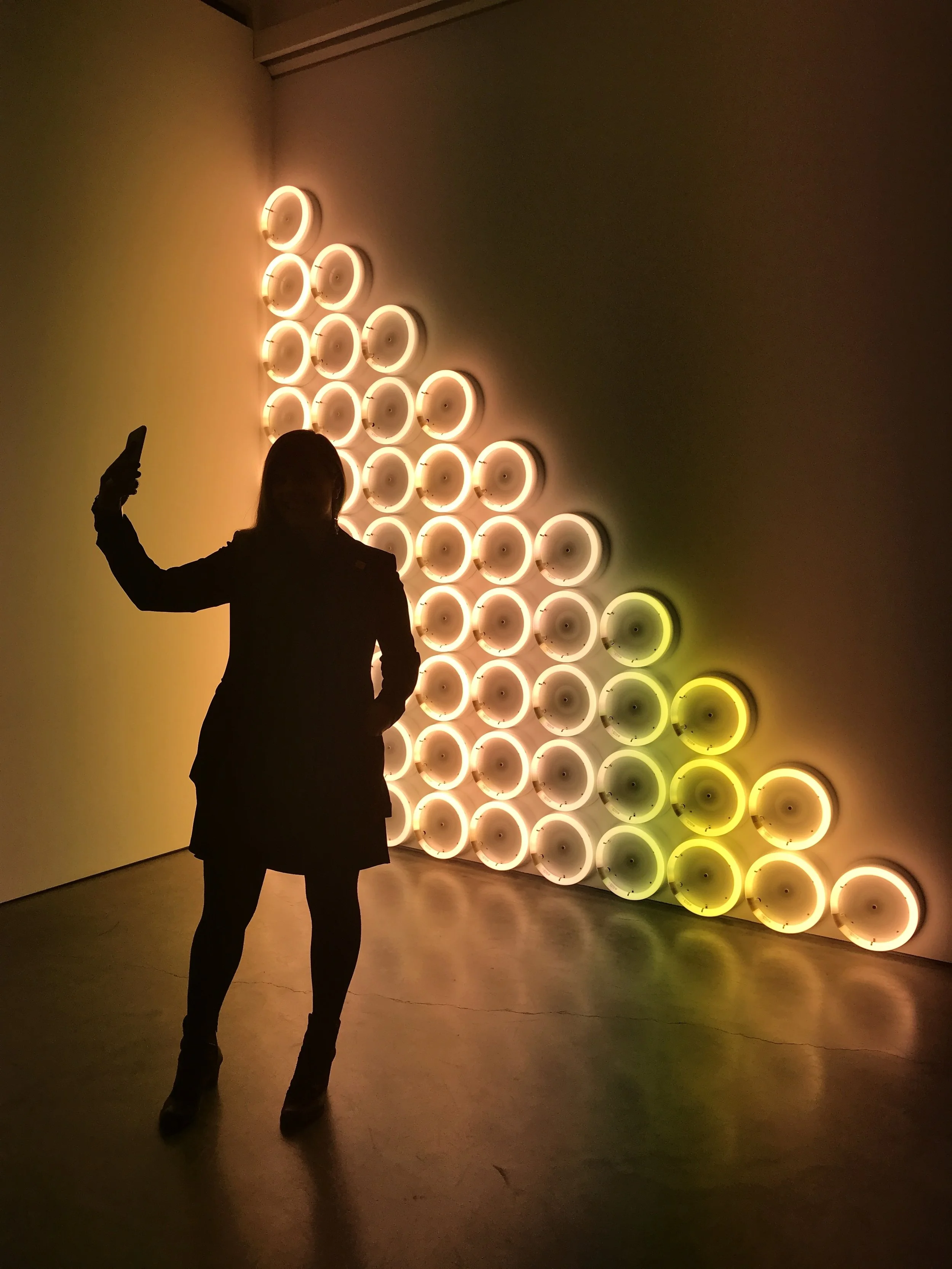 Silhouetted woman photographing a triangular wall installation of glowing amber and yellow circular lights.