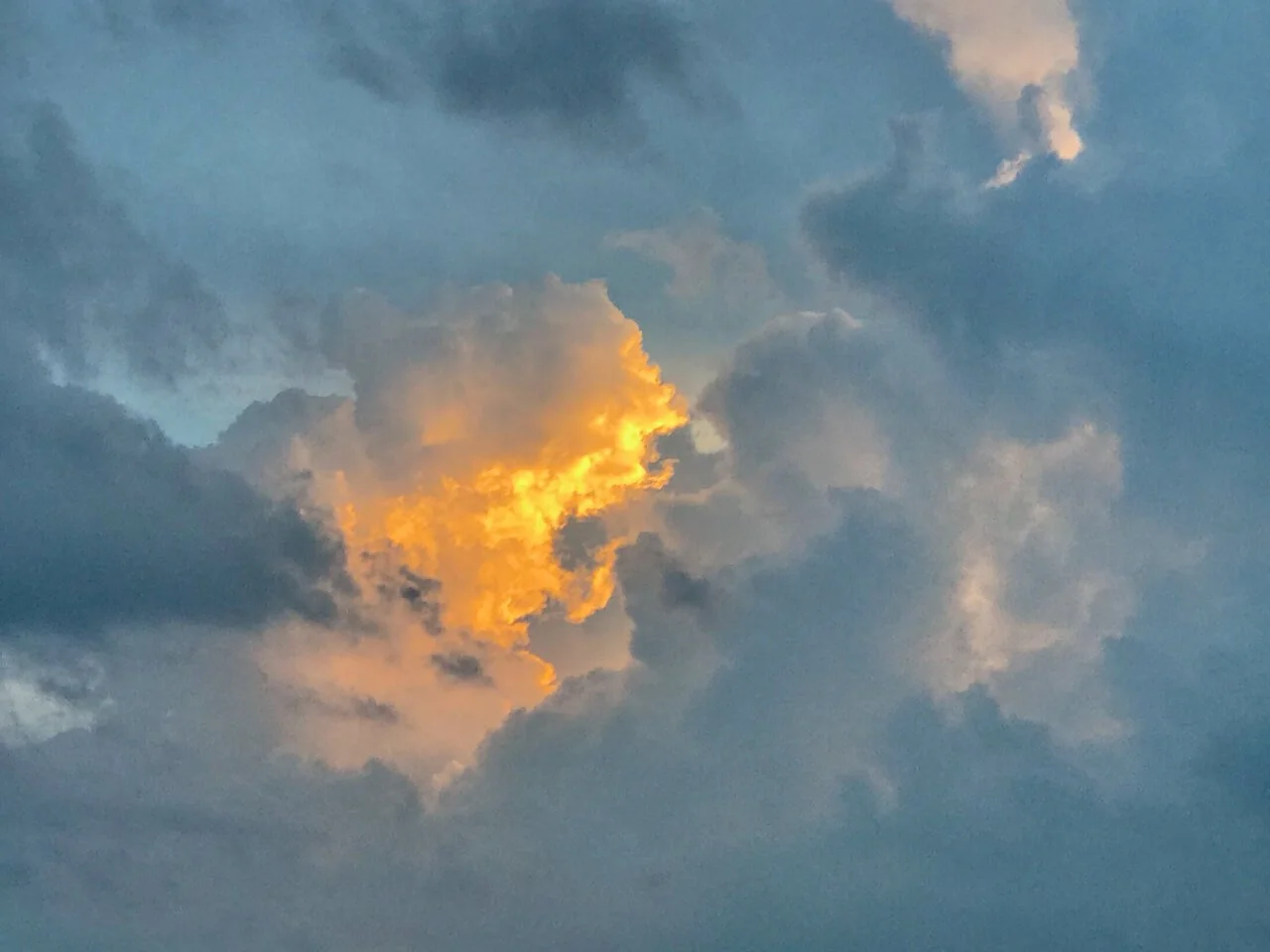 Billowing gray clouds rising up with blue sky in the background. In the center of the photo is a cloud reflecting the golden light of the sun.