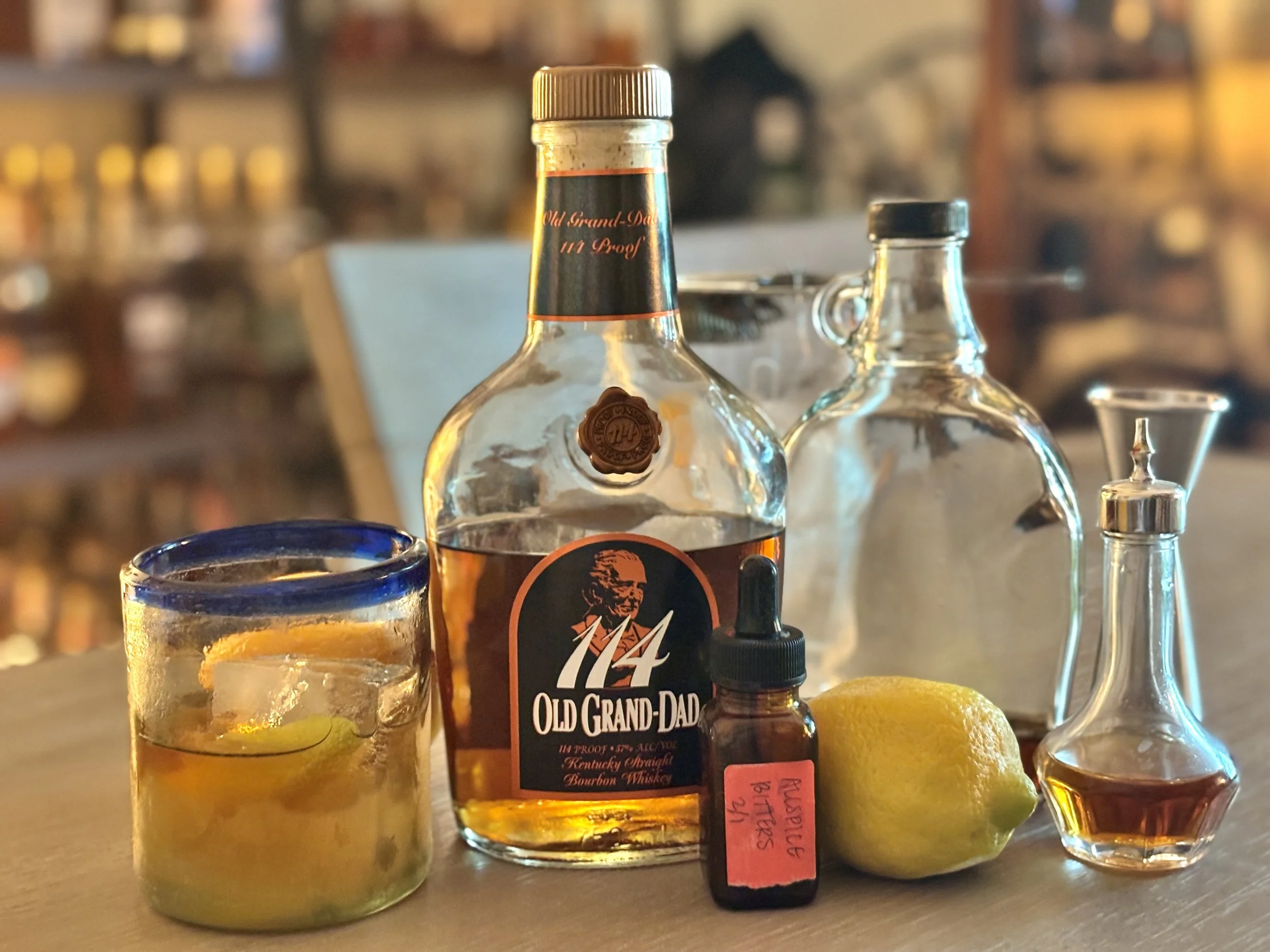 An old fashioned sits in a rocks glass to the left side of the phots with a bottle of OGD 114, a dropper bottle labeled Allspice bitters, a whole lemon, and a glass bottle of demerara syrup, and a dasher bottle of bitters then fill frame