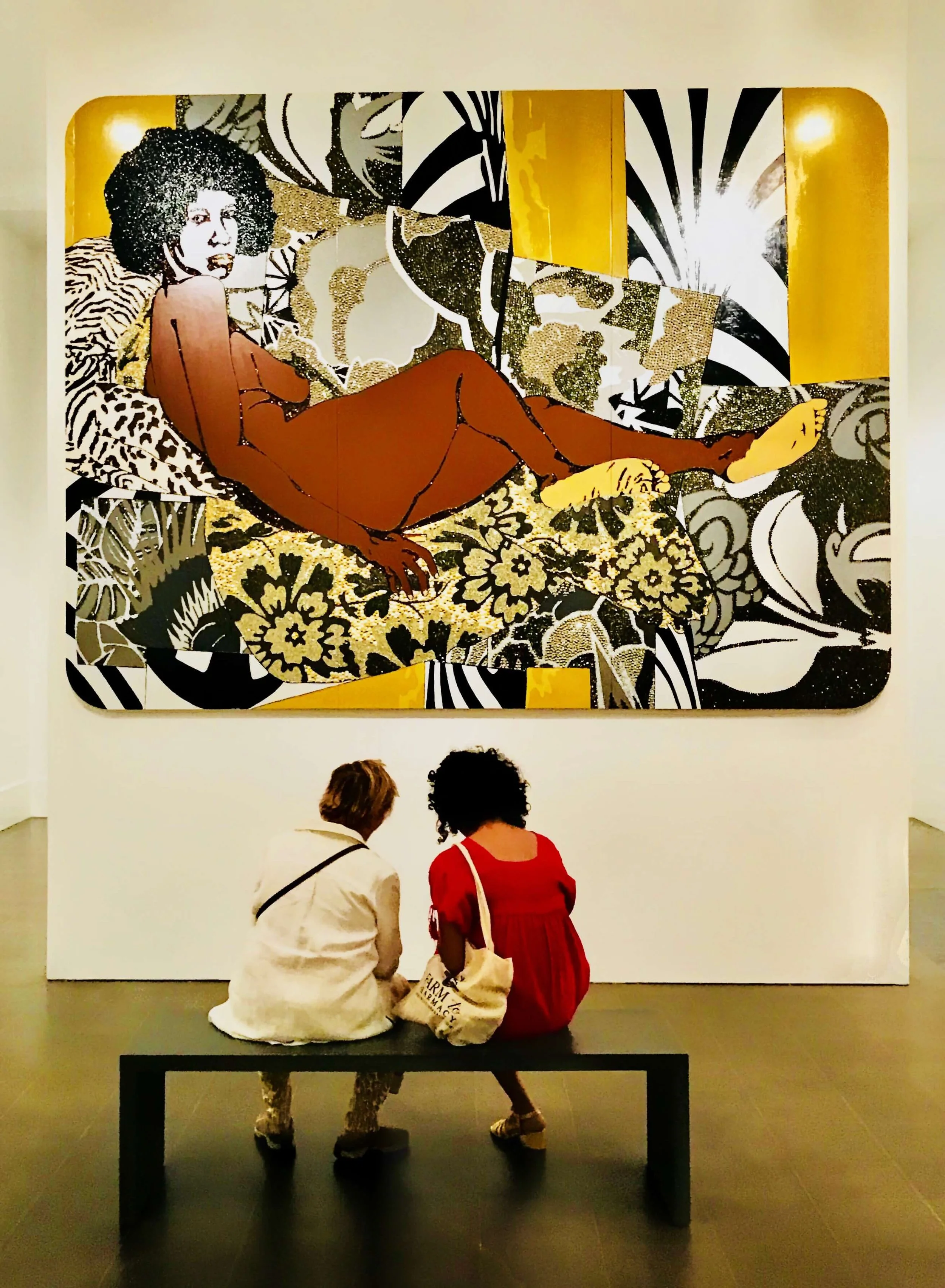 Two women sitting on a bench in front of a large painting by Mickalene Thomas called "A Little Taste Outside of Love. Taken at the Brooklyn Museum in Brooklyn, NY