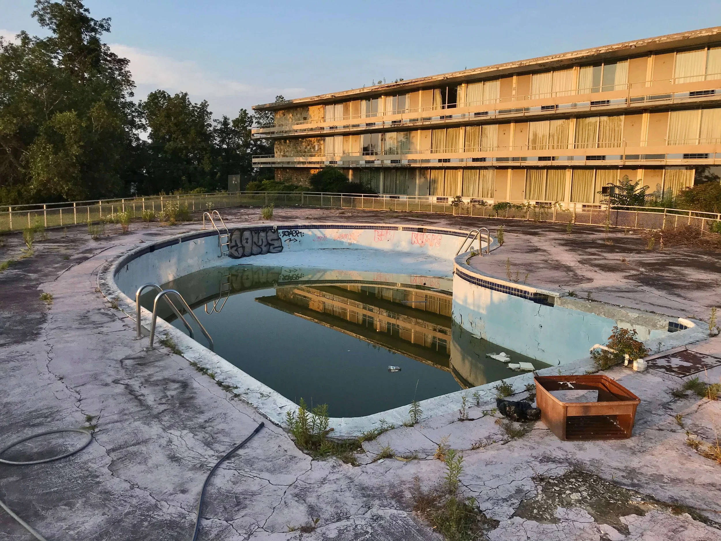 A color photograph taken at golden hour of an abandoned hotel in the background sitting in the glow of the sun.  In the foreground there is a half empty pool with discolored water and graffiti surrounded by cracked concrete with weeds growing up